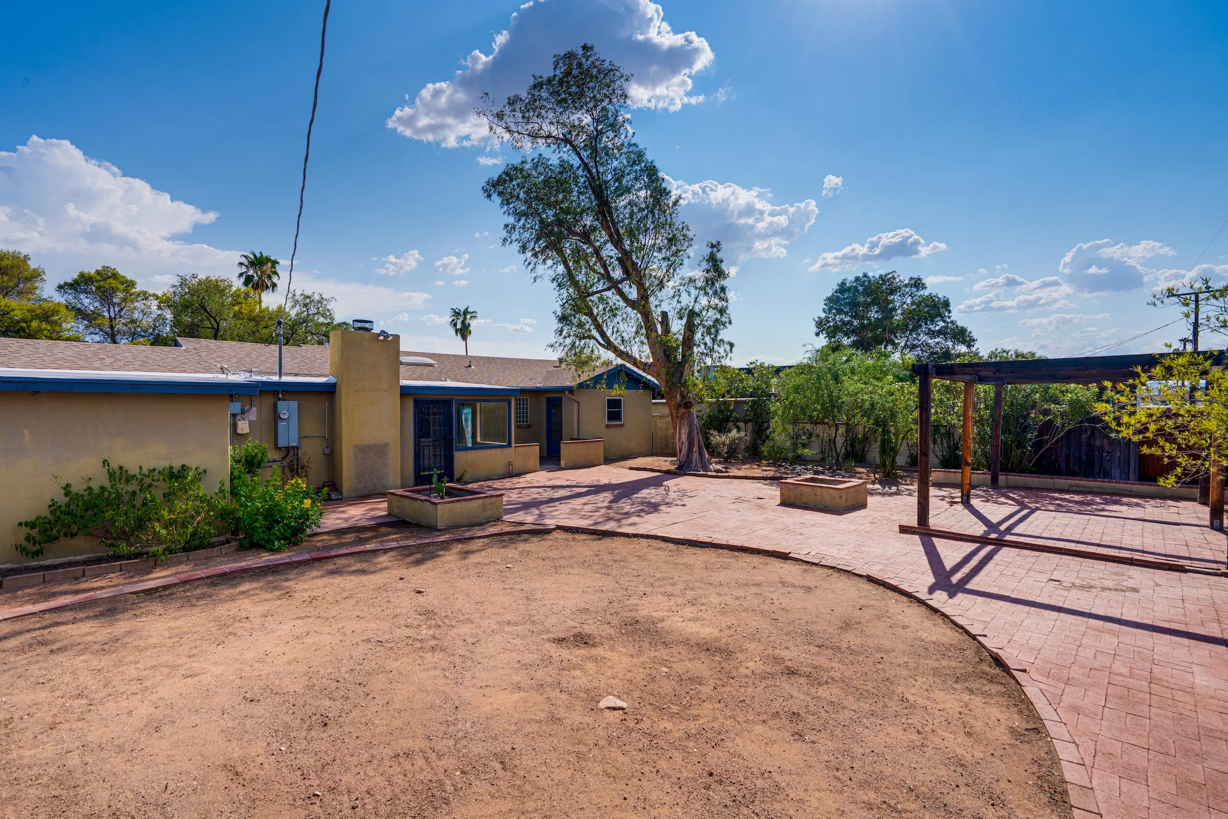 Backyard with a large tree, small garden beds, a house with a tiled roof, and a clear blue sky with scattered clouds.