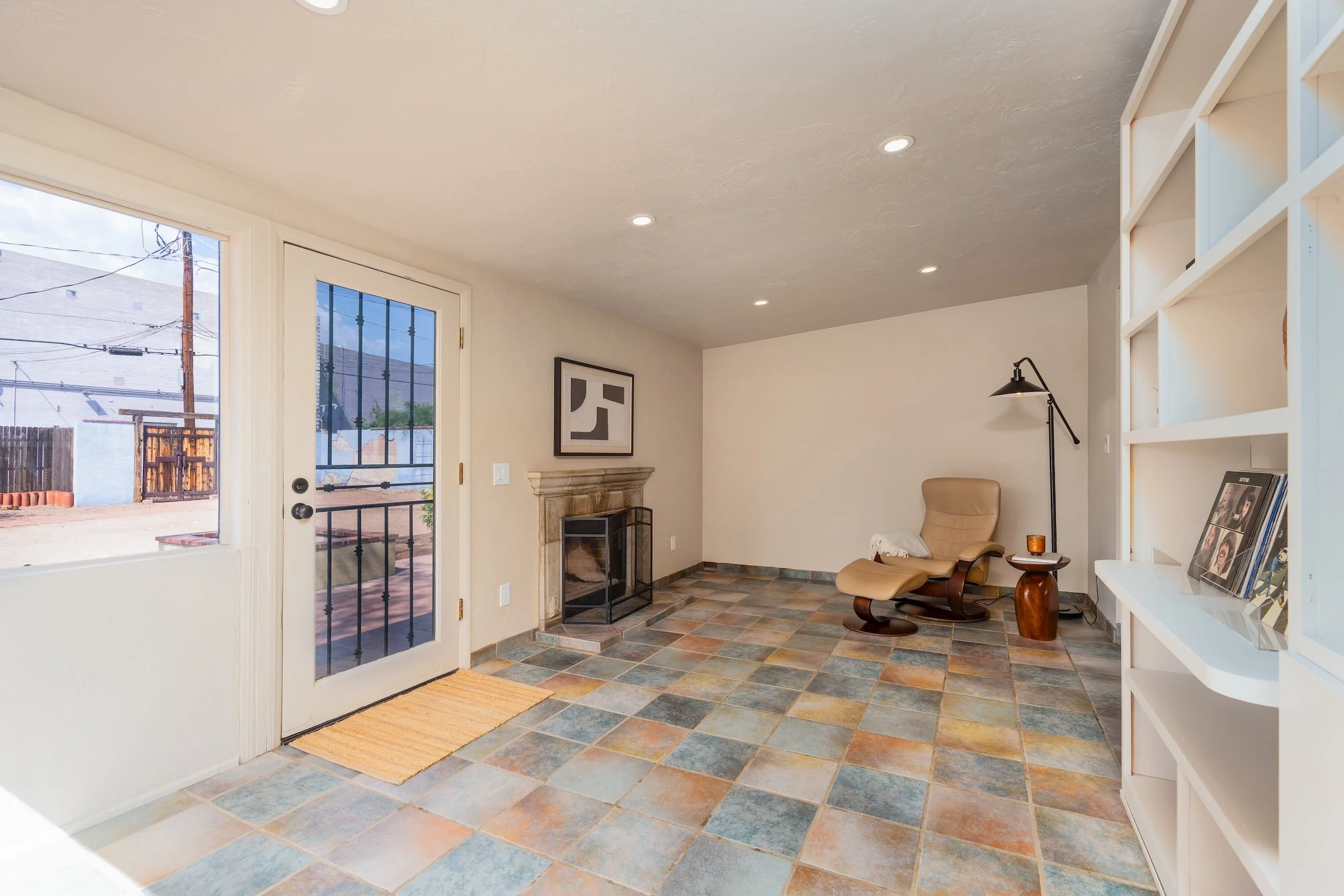 Living room with tiled floor, a beige recliner chair, a small side table with a candle, a floor lamp, a framed artwork on the wall, and a fireplace. There is a door with glass panels leading outside, with a view of a fence, utility pole, and neighbor