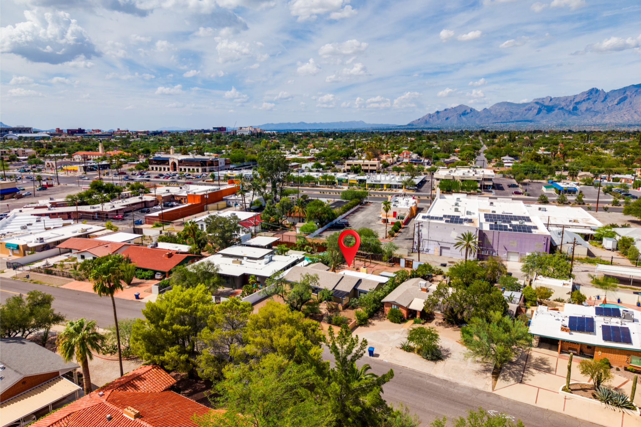 Aerial view of a city with green trees, low-rise buildings, and mountains in the background under a partly cloudy sky.