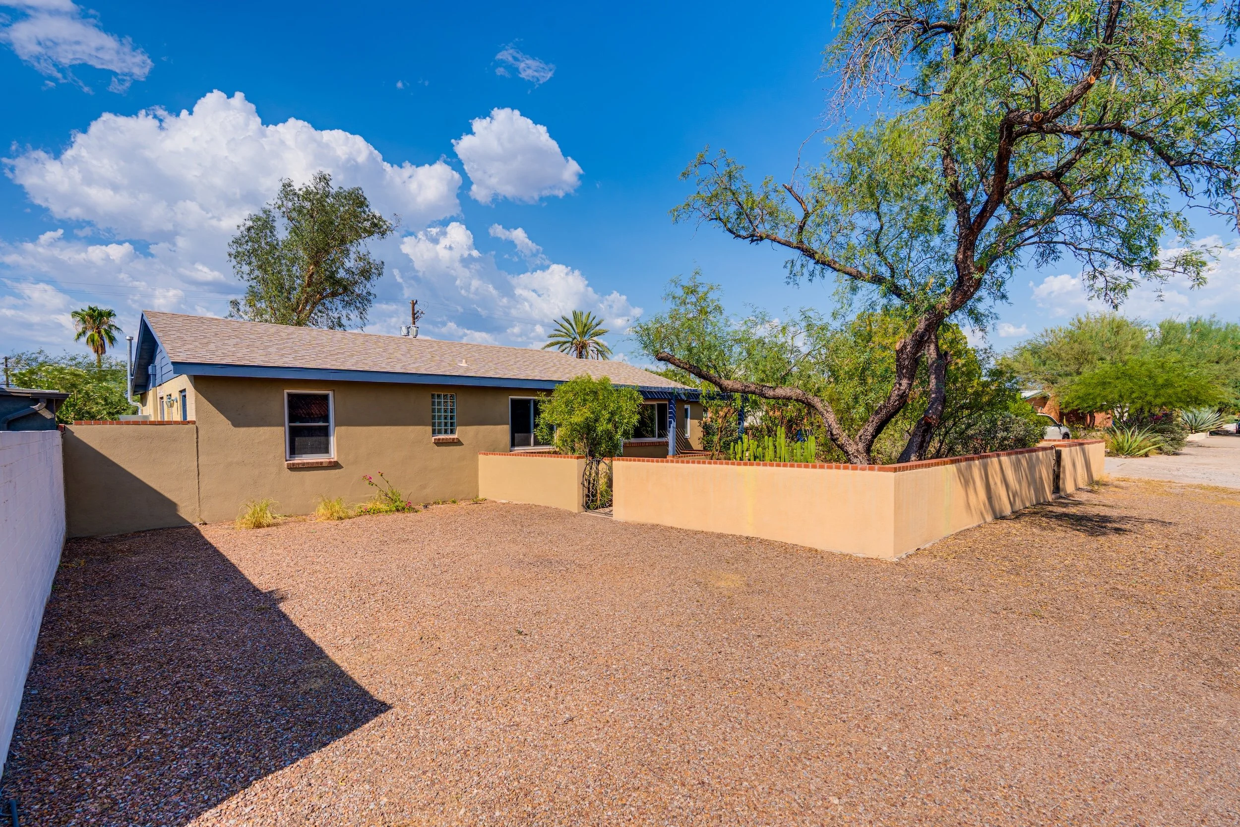 A backyard with a gravel ground, a beige house with small windows, and tall green trees against a blue sky with scattered white clouds.