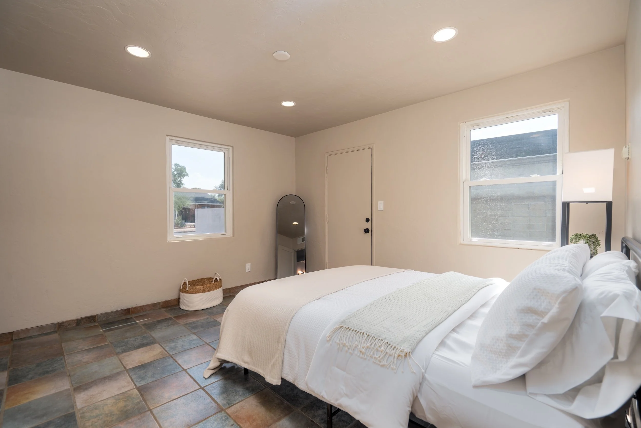 A bedroom featuring a bed with white bedding, two windows allowing natural light, a mirror, and a basket on a tiled floor.