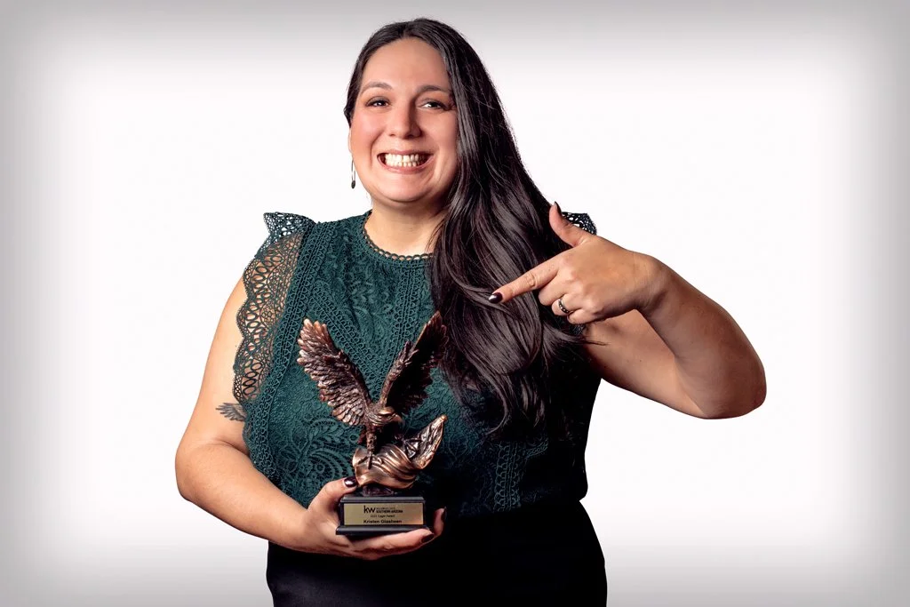 Woman with long dark hair smiling and pointing to an award she is holding, which features an eagle with outstretched wings, against a plain white background.