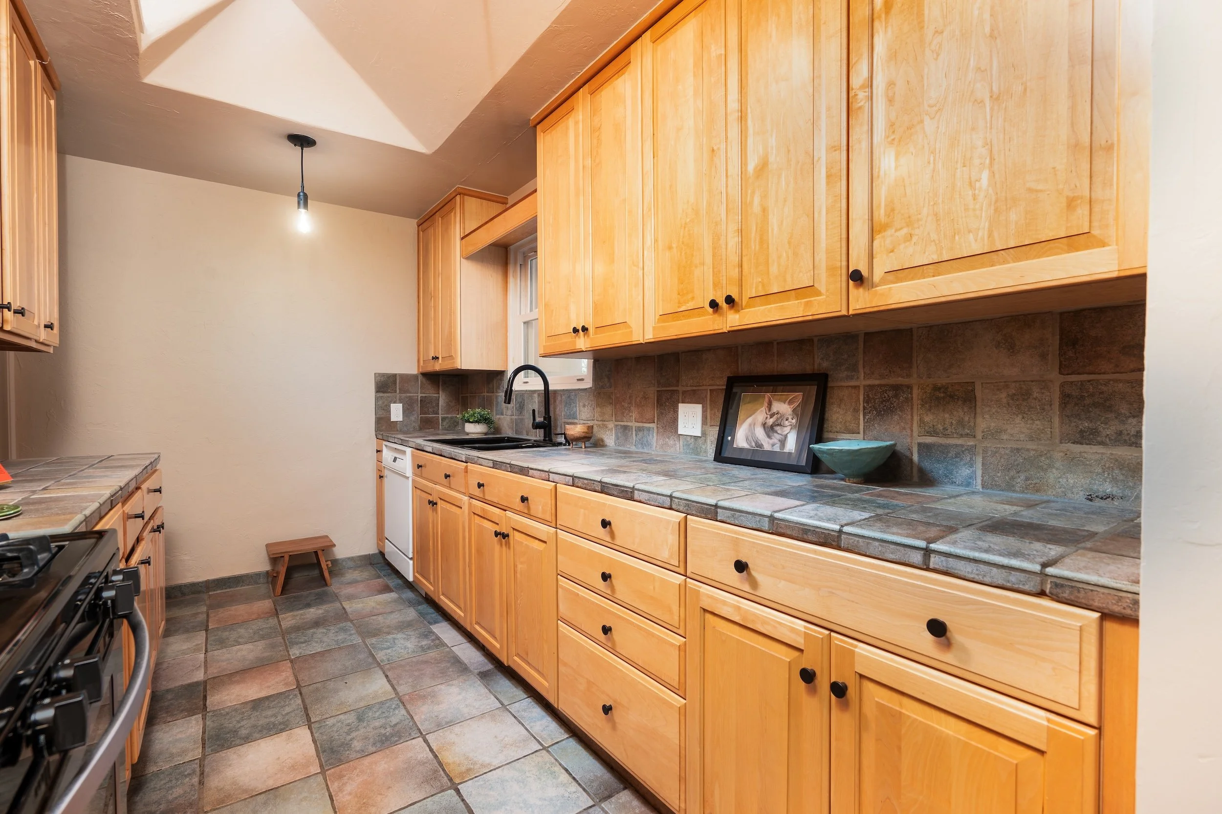 A kitchen with wooden cabinets, a tiled countertop, and floor. There is a framed cat picture and a bowl on the counter, a small plant, and a window above the sink. An empty stool is near the wall, and a hanging light fixture is above.
