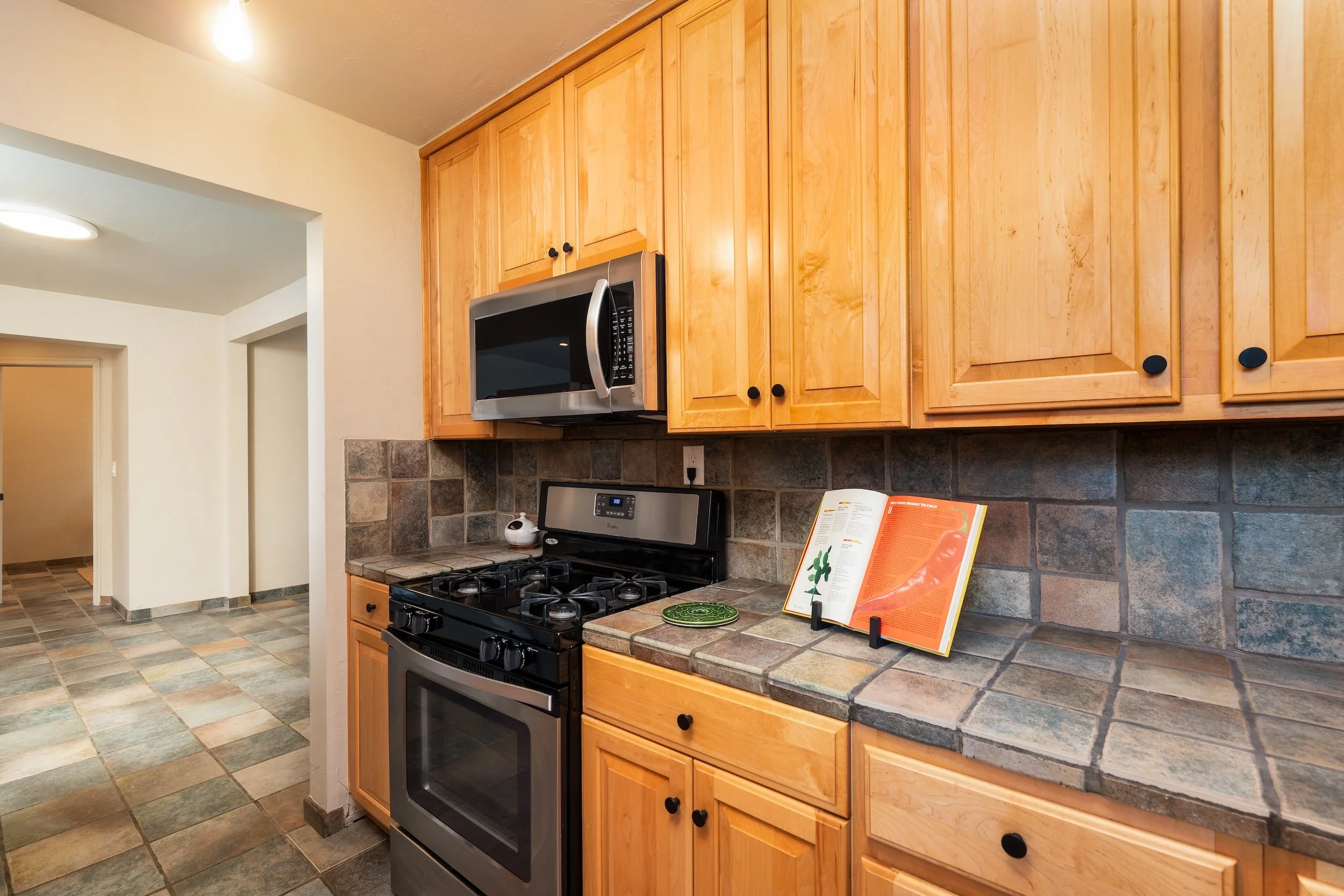 A kitchen with wooden cabinets, a gas stove, a microwave above the stove, a tiled backsplash, and an open cookbook on a stand.