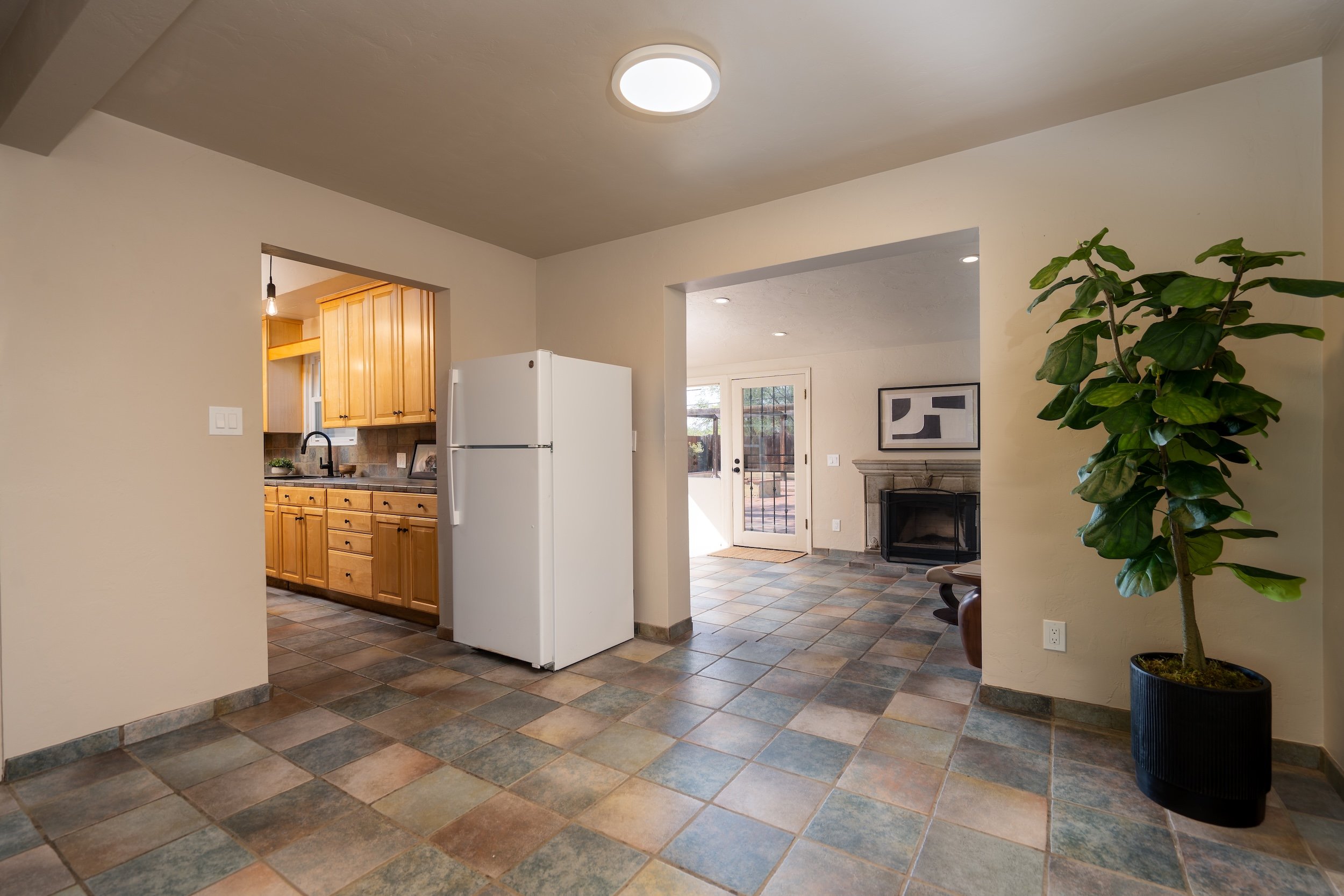 Interior view of a house with a tiled floor, a white refrigerator, a kitchen with wooden cabinets, a fireplace, and a large houseplant in a black pot near the wall.