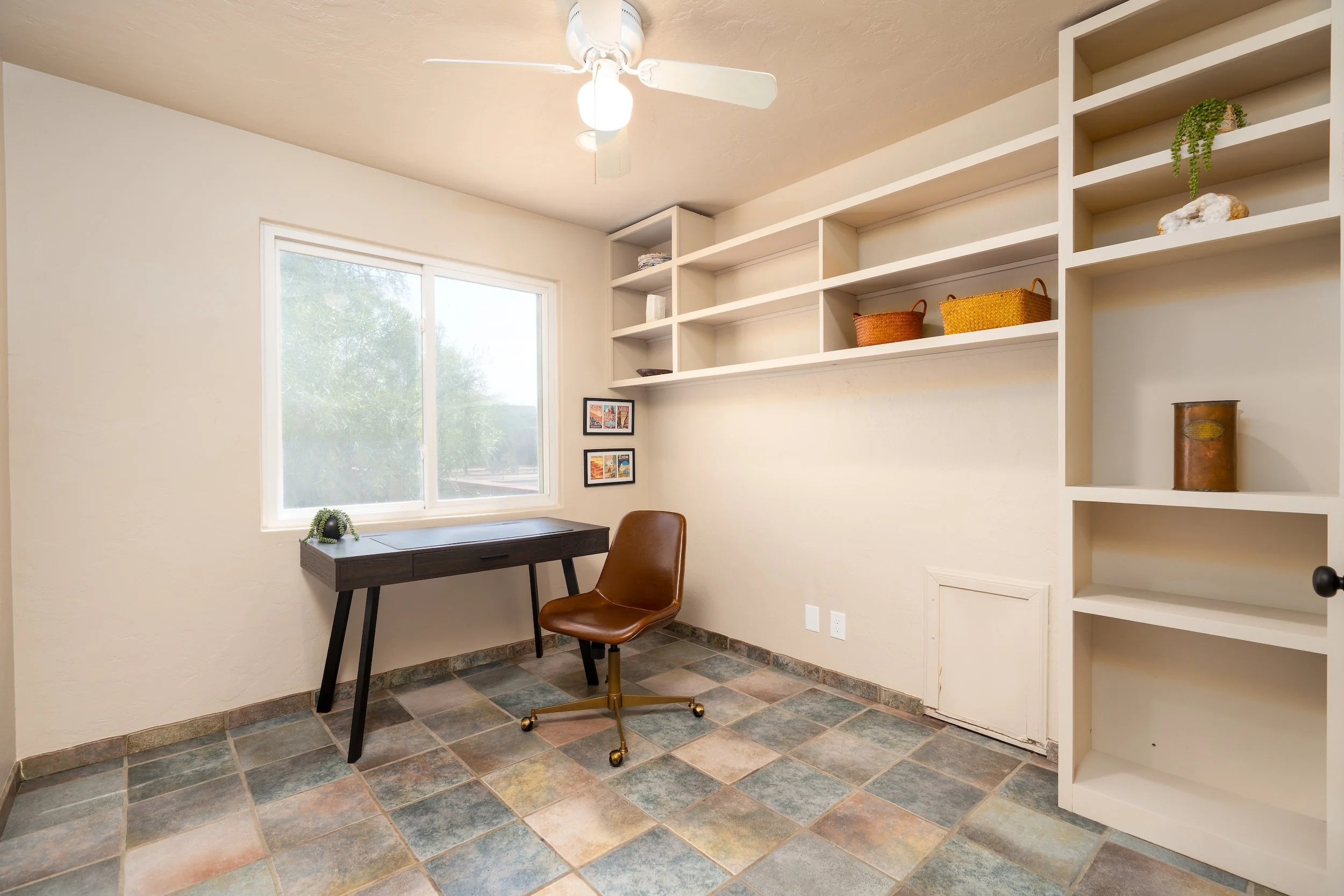 A small home office with a window, a dark wooden desk, a brown office chair, white built-in shelving with decorative baskets, a potted plant, and framed pictures on the wall.