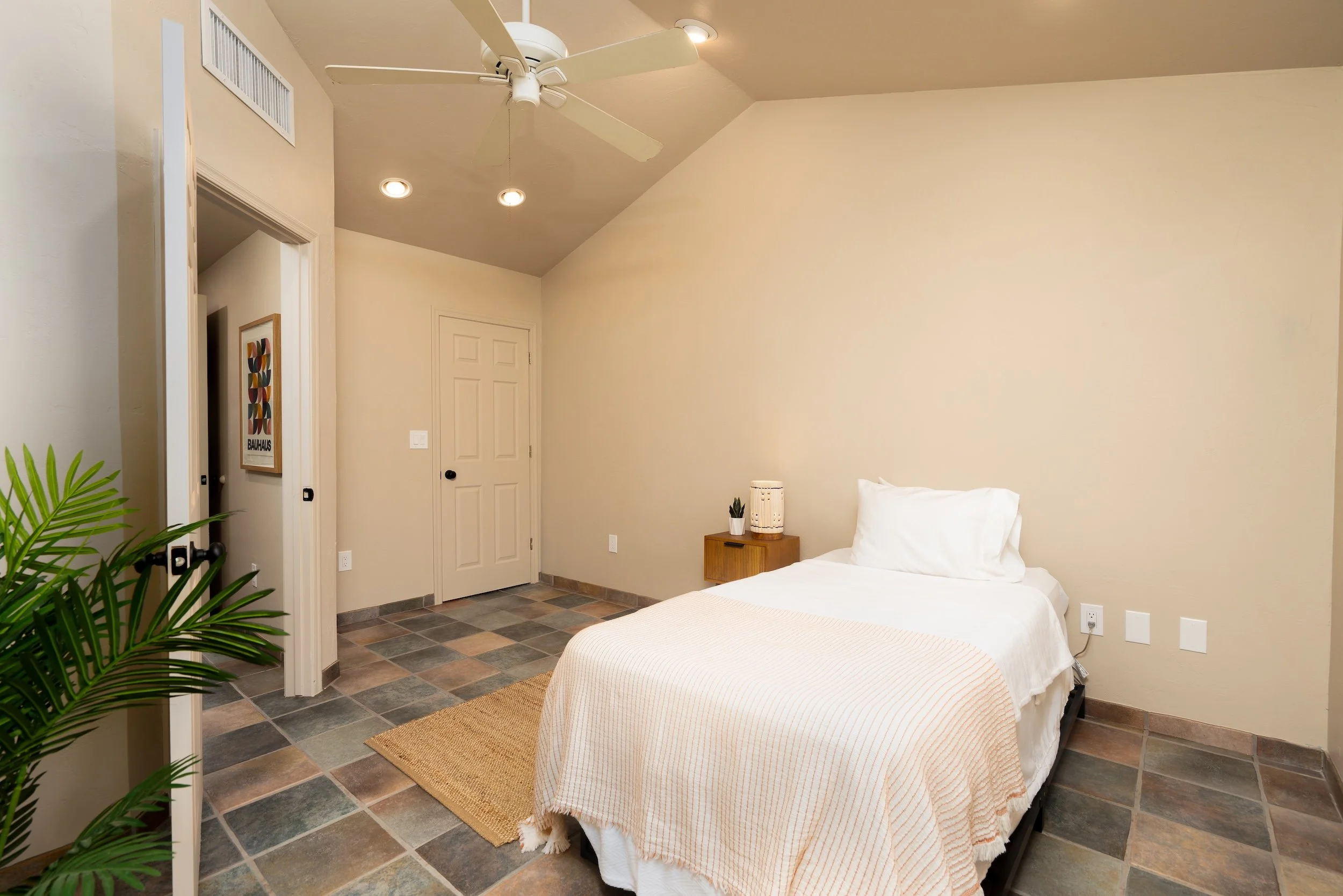 A bedroom with a bed covered in a light-colored blanket and a white pillow. There are small decor items on a brown nightstand next to the bed. The room has beige walls, a tiled floor, a ceiling fan, and recessed lighting. A door and some artwork are 