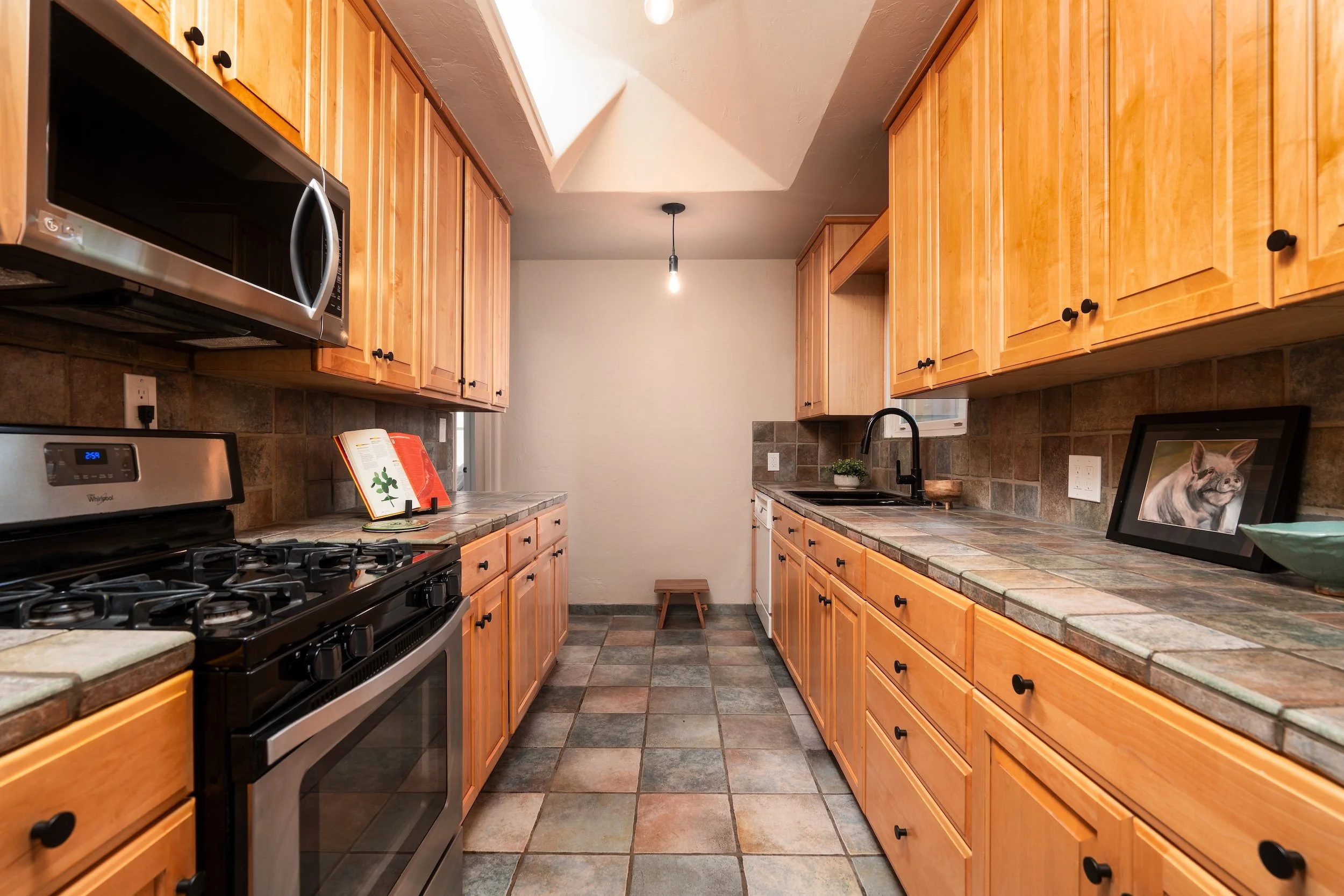 Kitchen with wood cabinets, tiled counters and backsplash, stainless steel stove and microwave, a small plant, framed photo of a dog, open cookbook, and a stool under the ceiling light.