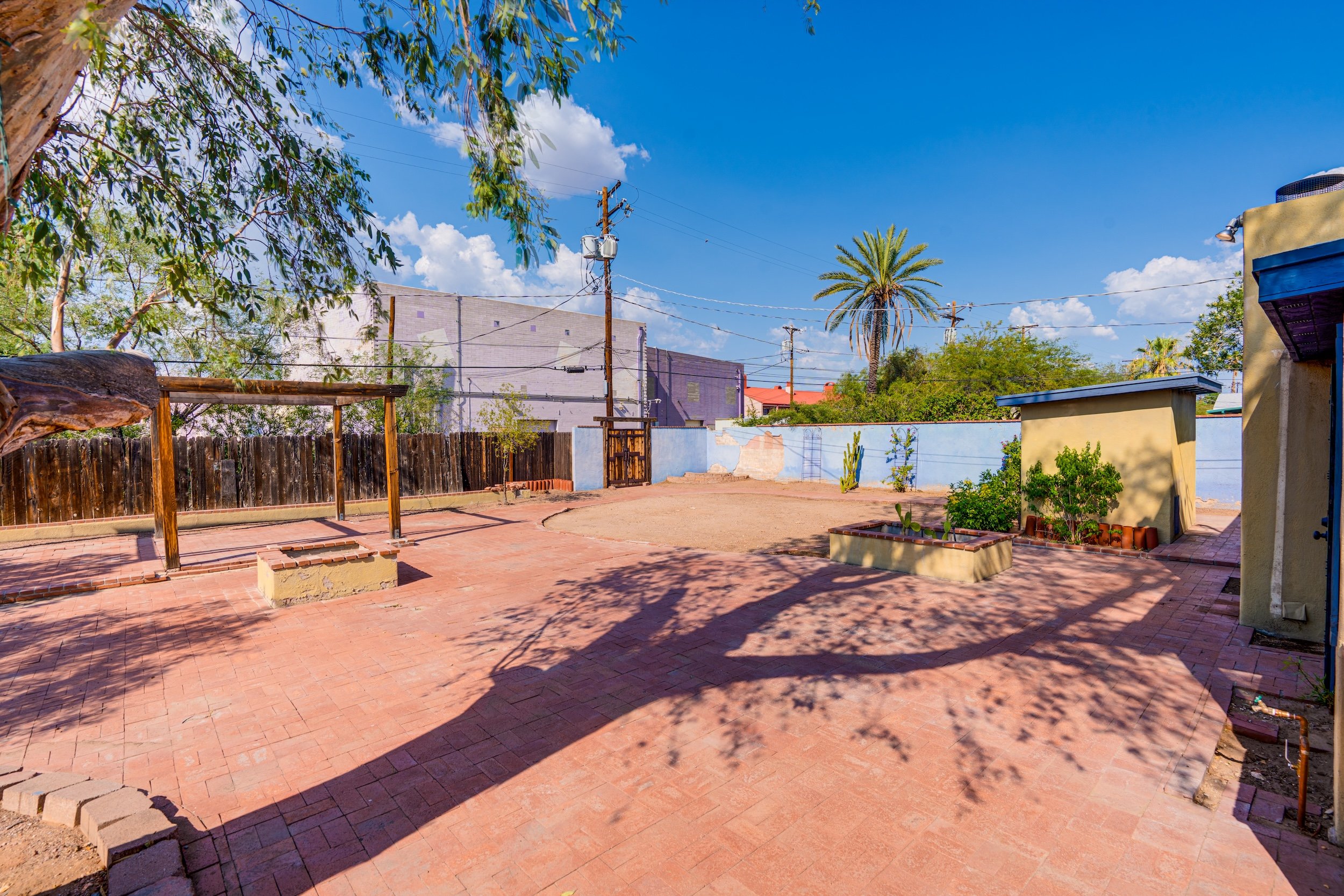 A backyard patio with red brick paving, small raised garden beds with plants, a wooden pergola frame, a tall palm tree, a blue sky with clouds, and a fence.