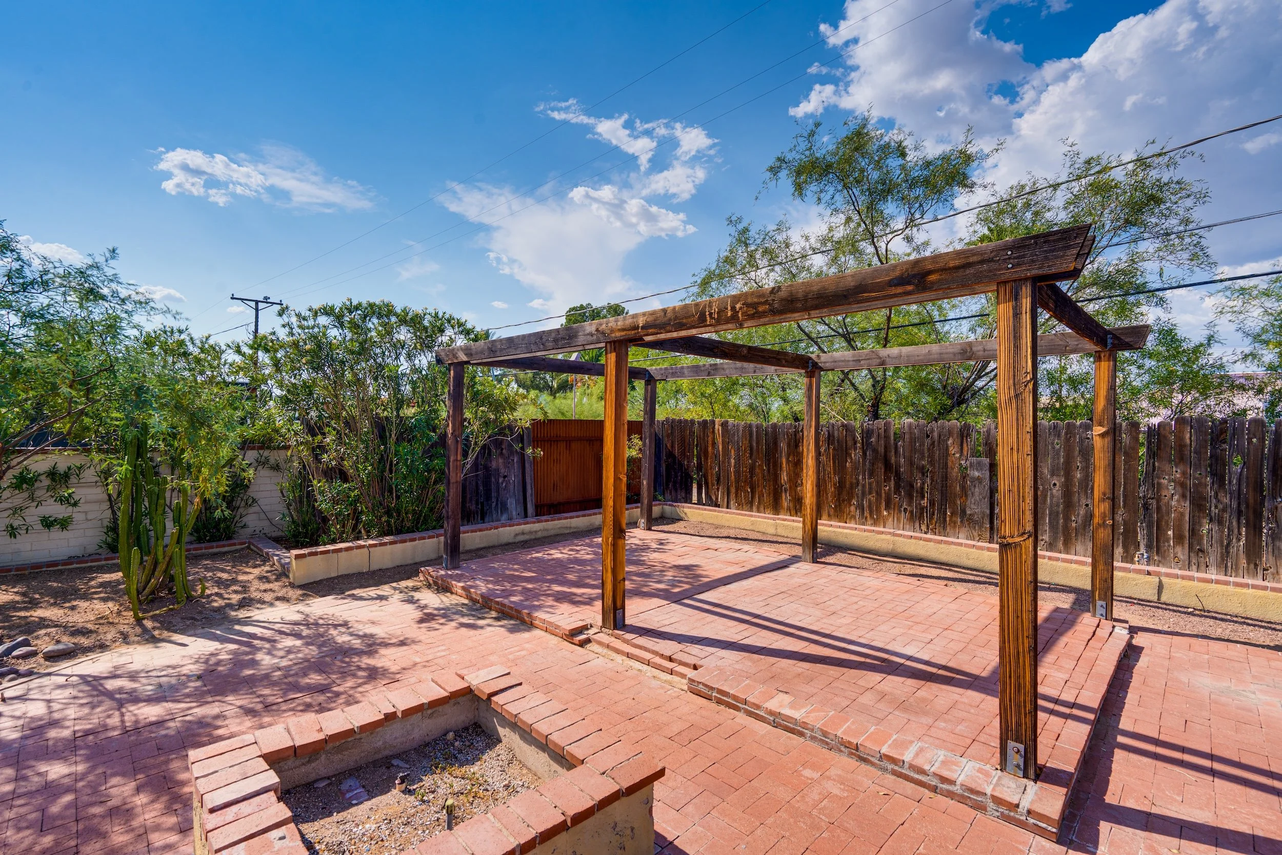 An unfinished outdoor structure with wooden posts and crossbeams over a brick patio, surrounded by a wooden fence and plants, under a bright blue sky with clouds.