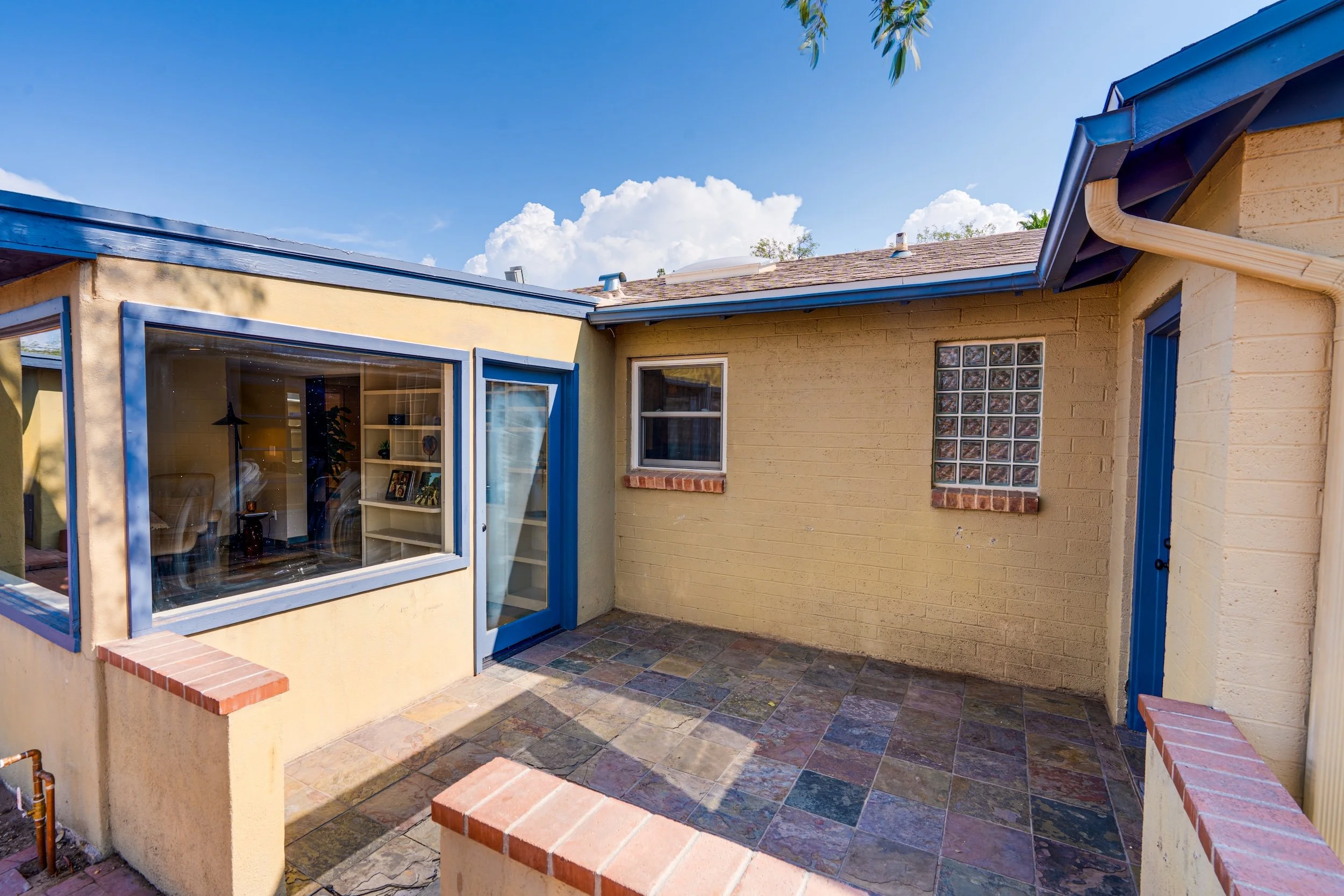 A small outdoor patio area in the backyard of a house with beige walls, blue window and door frames, a glass block window, a roof with shingles, and a cloudy blue sky overhead.