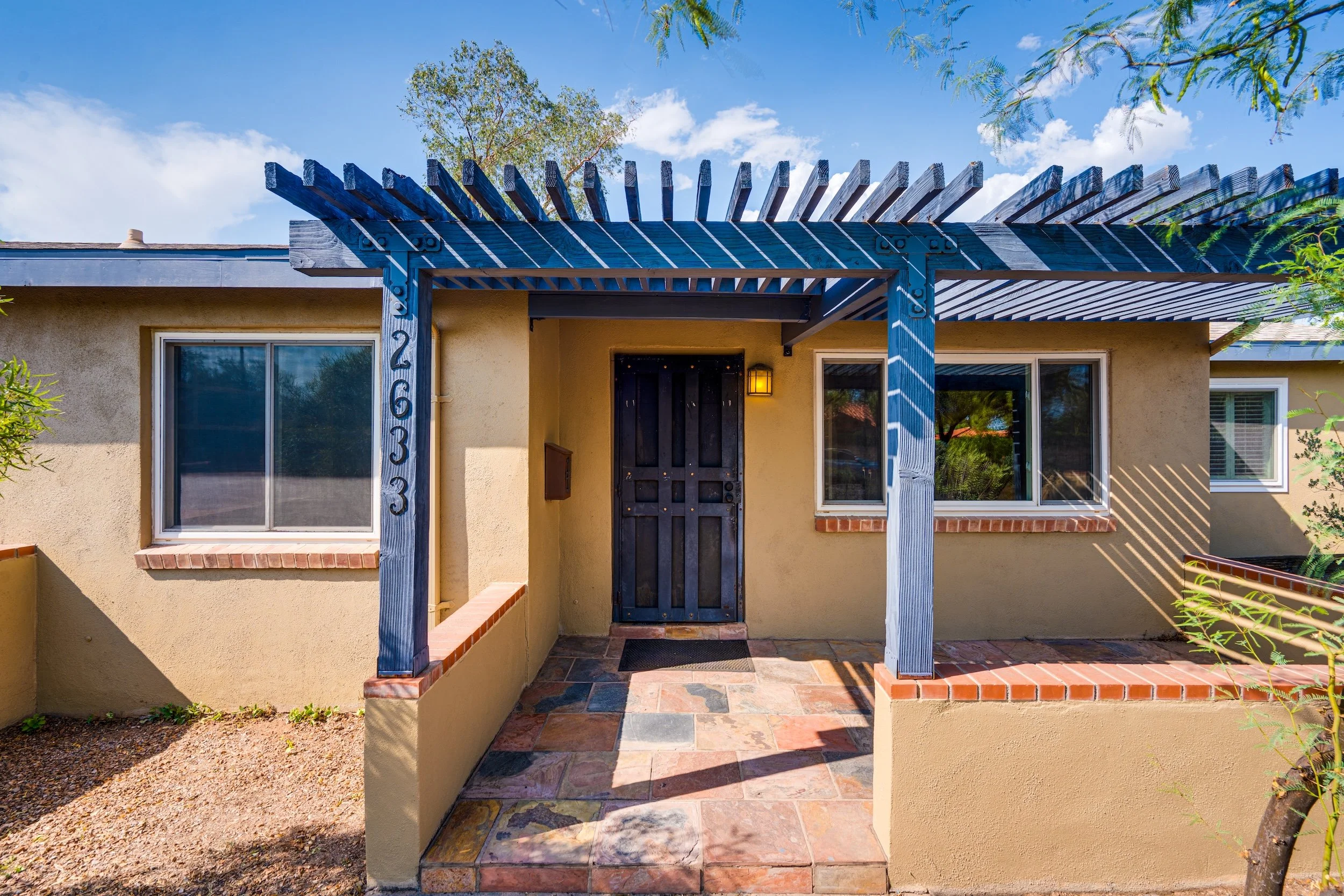 Front view of a house with a beige exterior, black front door, and a blue wooden pergola above the entrance. There are two large windows on either side of the door, with brick window sills. The walkway and steps leading to the door are made of colorf