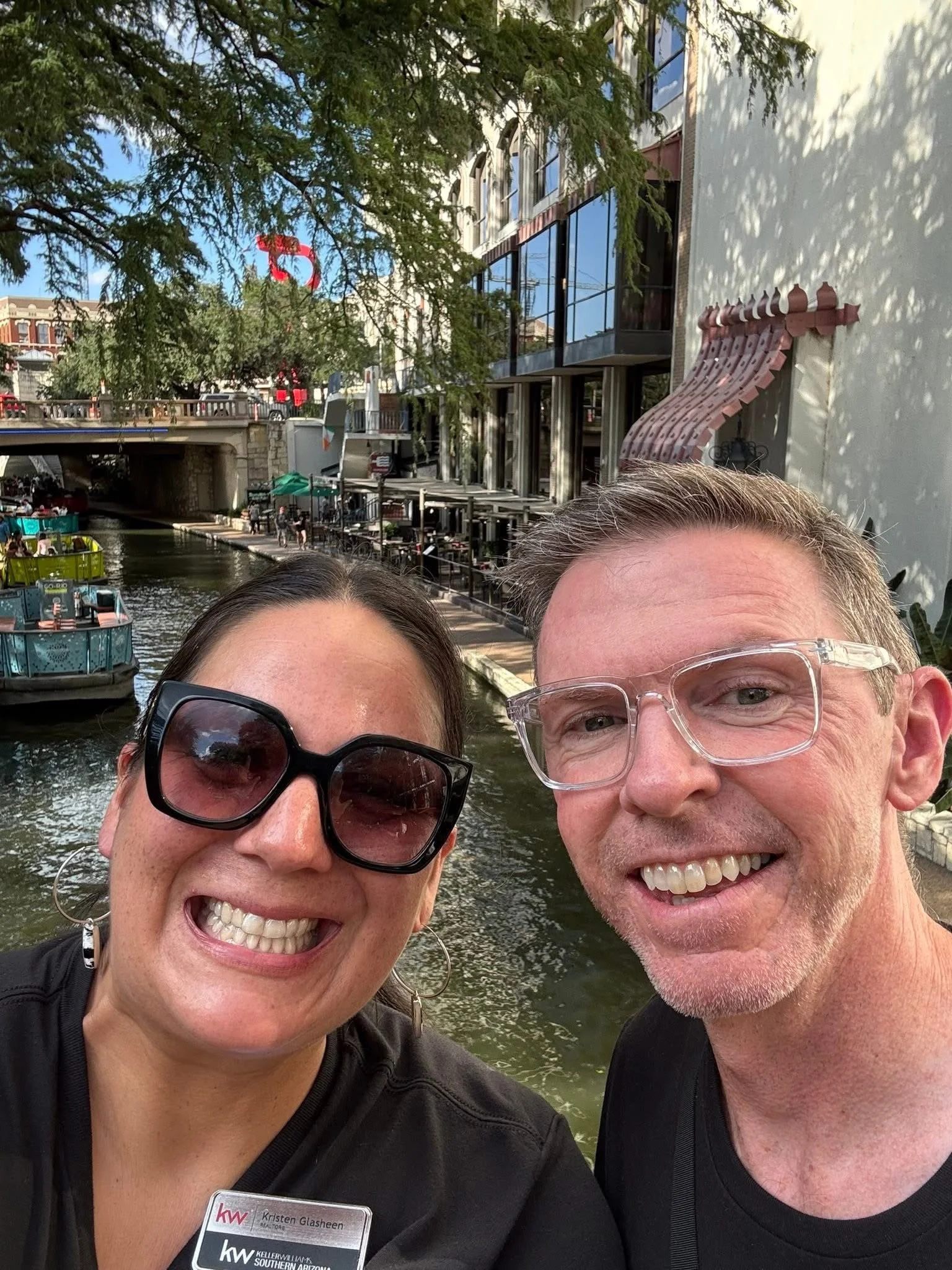 A smiling woman and man taking a selfie by a canal with boats, along a walkway lined with trees and buildings, on a sunny day.