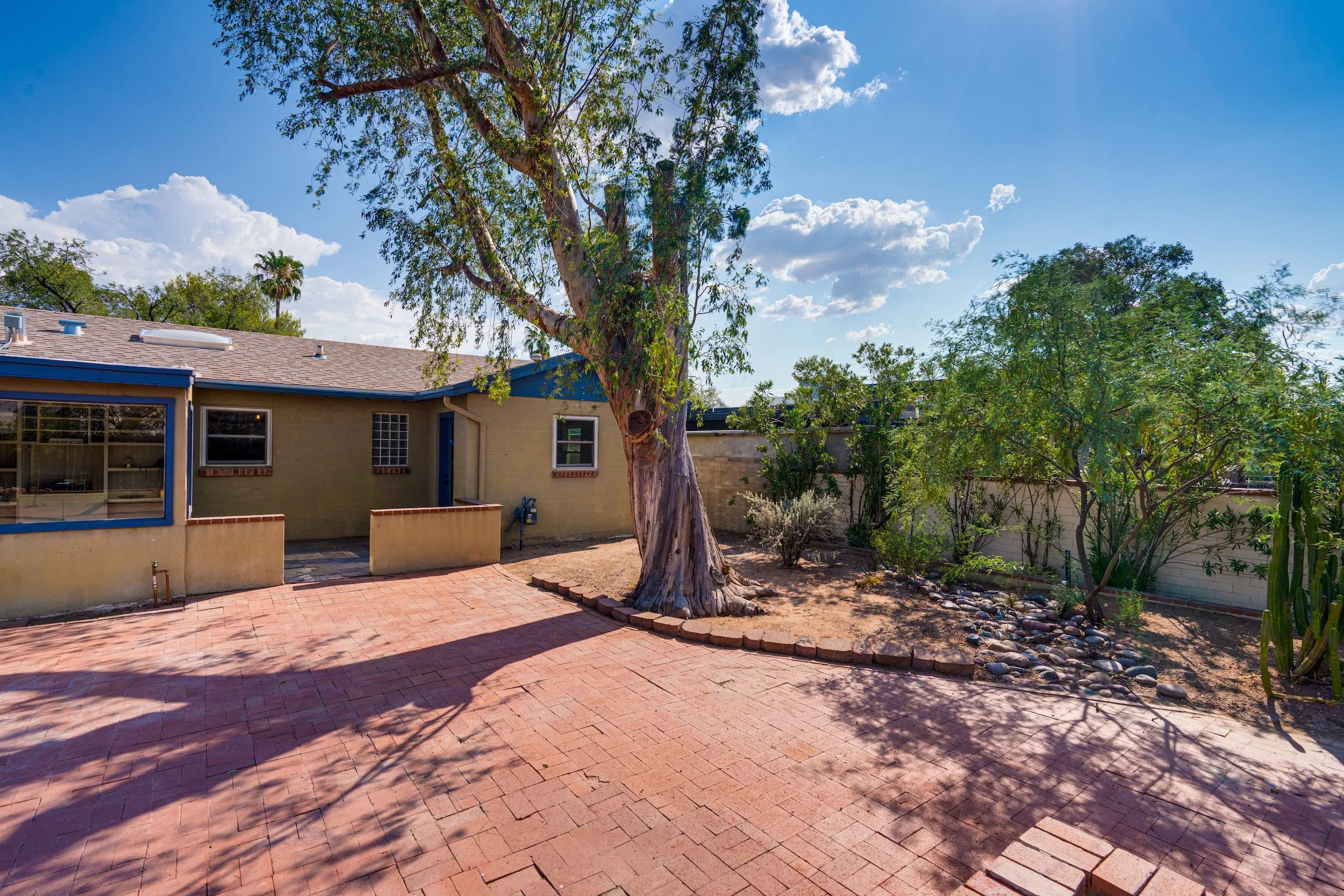 A backyard with a large tree, a brick-paved patio area, and a house with a yellow exterior and blue trim. The sky is blue with some scattered clouds.