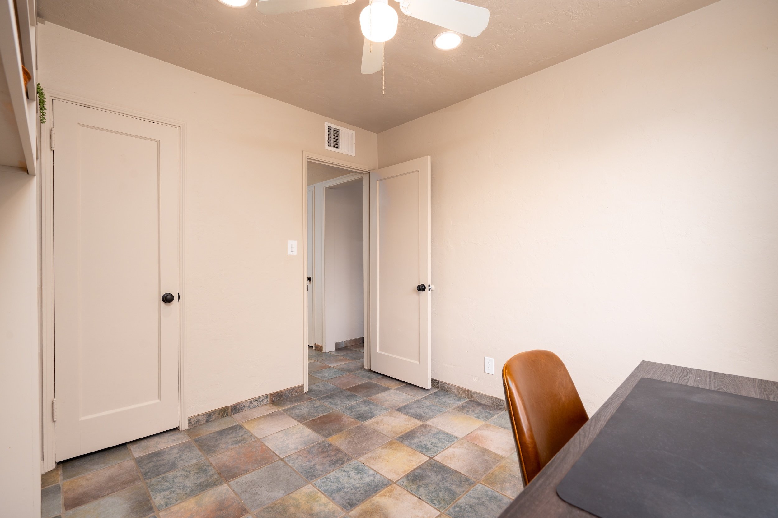 Interior of a room with tiled floor, white walls, open door leading to a hallway, ceiling fan with lights, partial view of a table and brown chair.