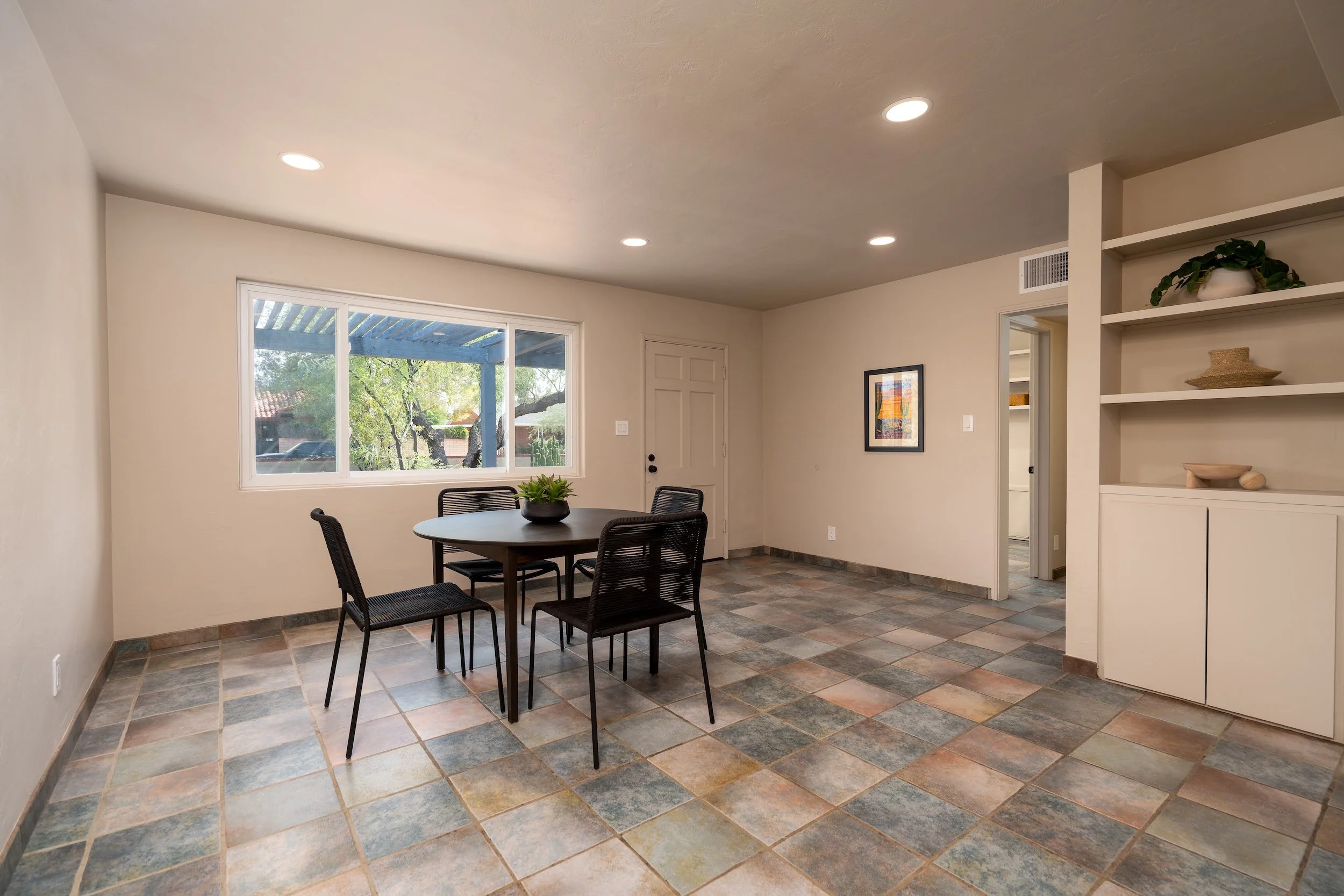 Interior of a dining room with a round black table, four black chairs, a window showing trees outside, a framed picture on the wall, built-in shelves with decorative items, and tiled flooring.
