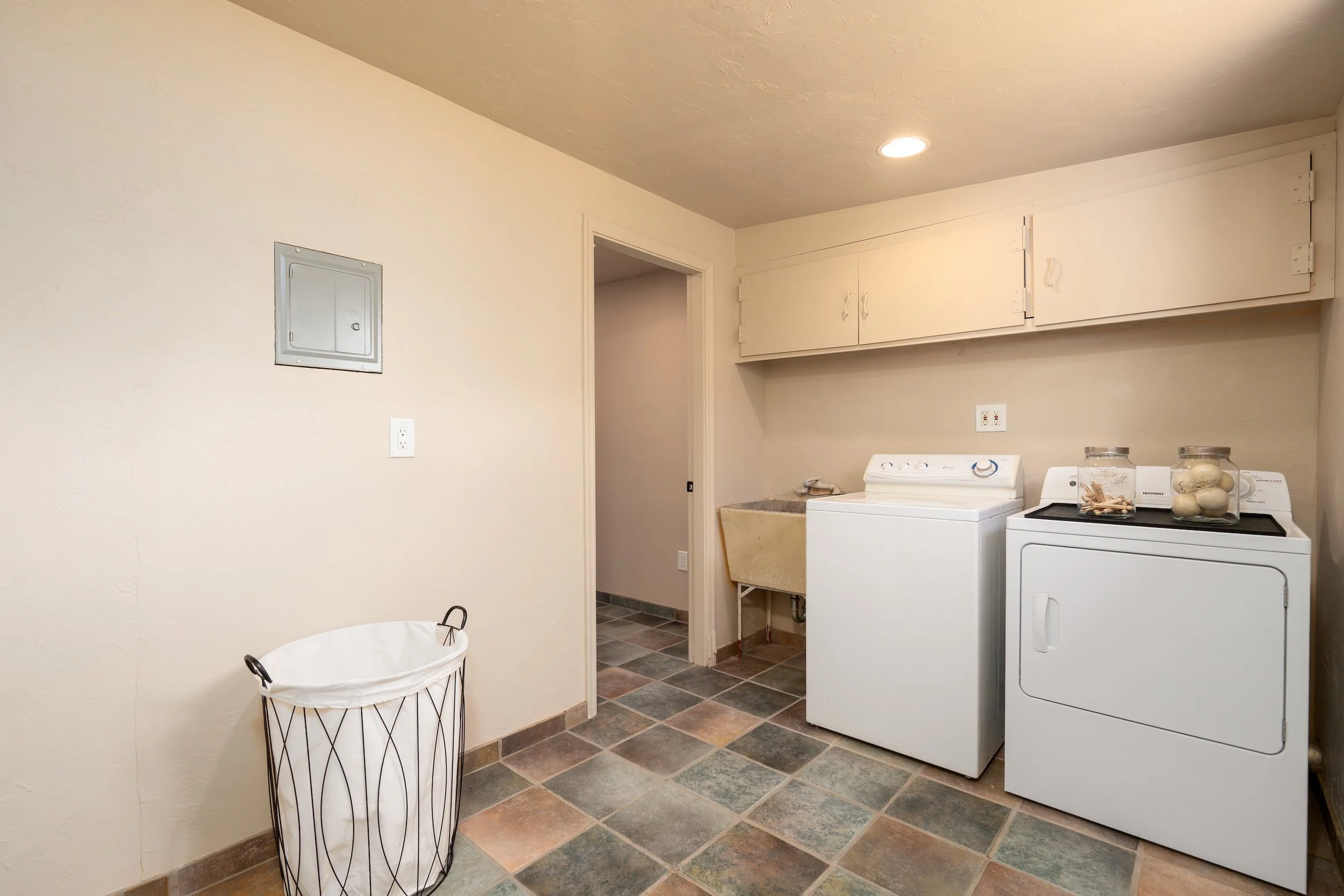 Laundry room with washing machine and dryer, laundry basket, wall cabinets, electrical panel, and tiled floor.