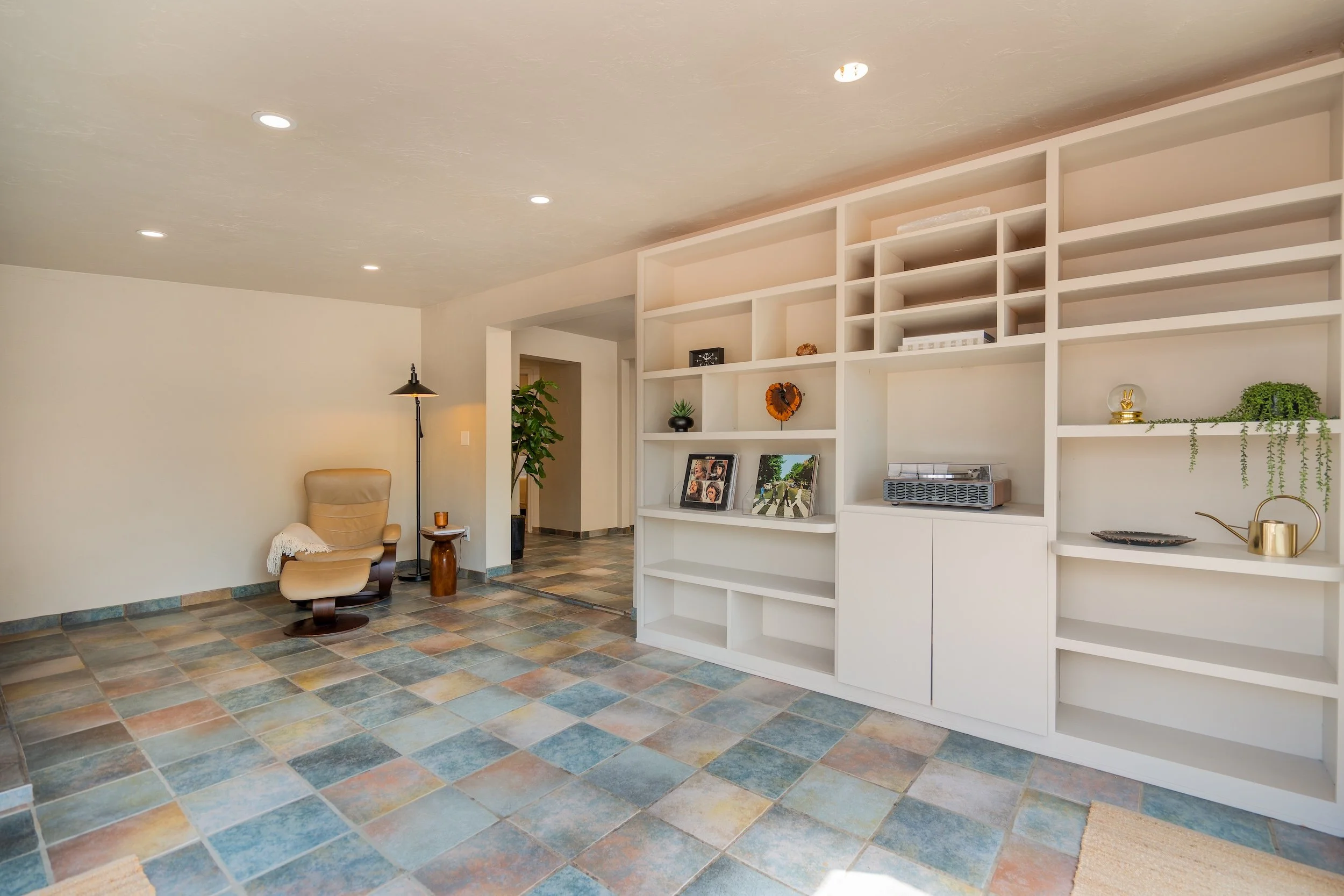 Living room with built-in white shelving, beige armchair, potted plants, pictures, and decorative items, with a tiled floor and ceiling lights.