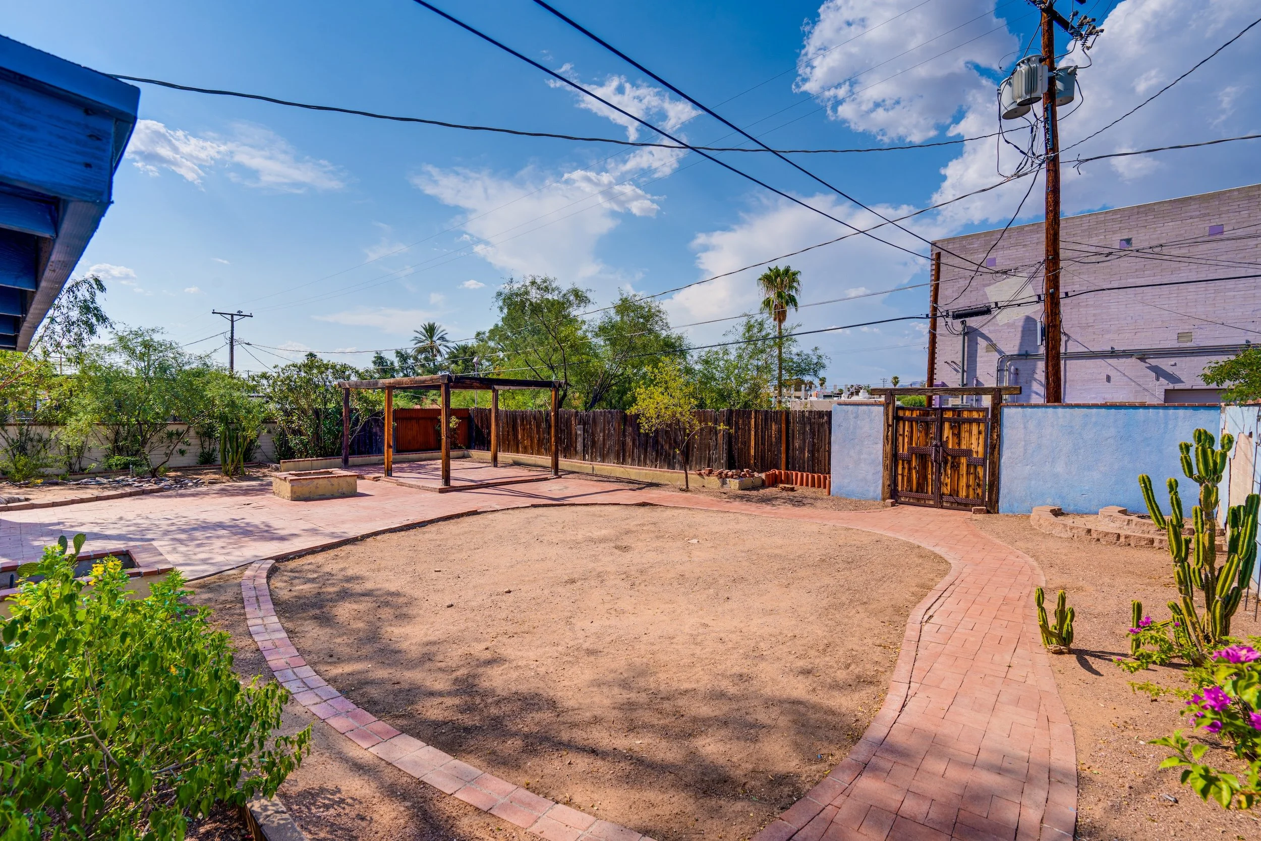 An outdoor backyard with a curved brick pathway, a wooden pergola, green bushes, flowering plants, a tall cactus, trees, a wooden and a chain link fence, utility poles and wires, a white building, and a partly cloudy blue sky.
