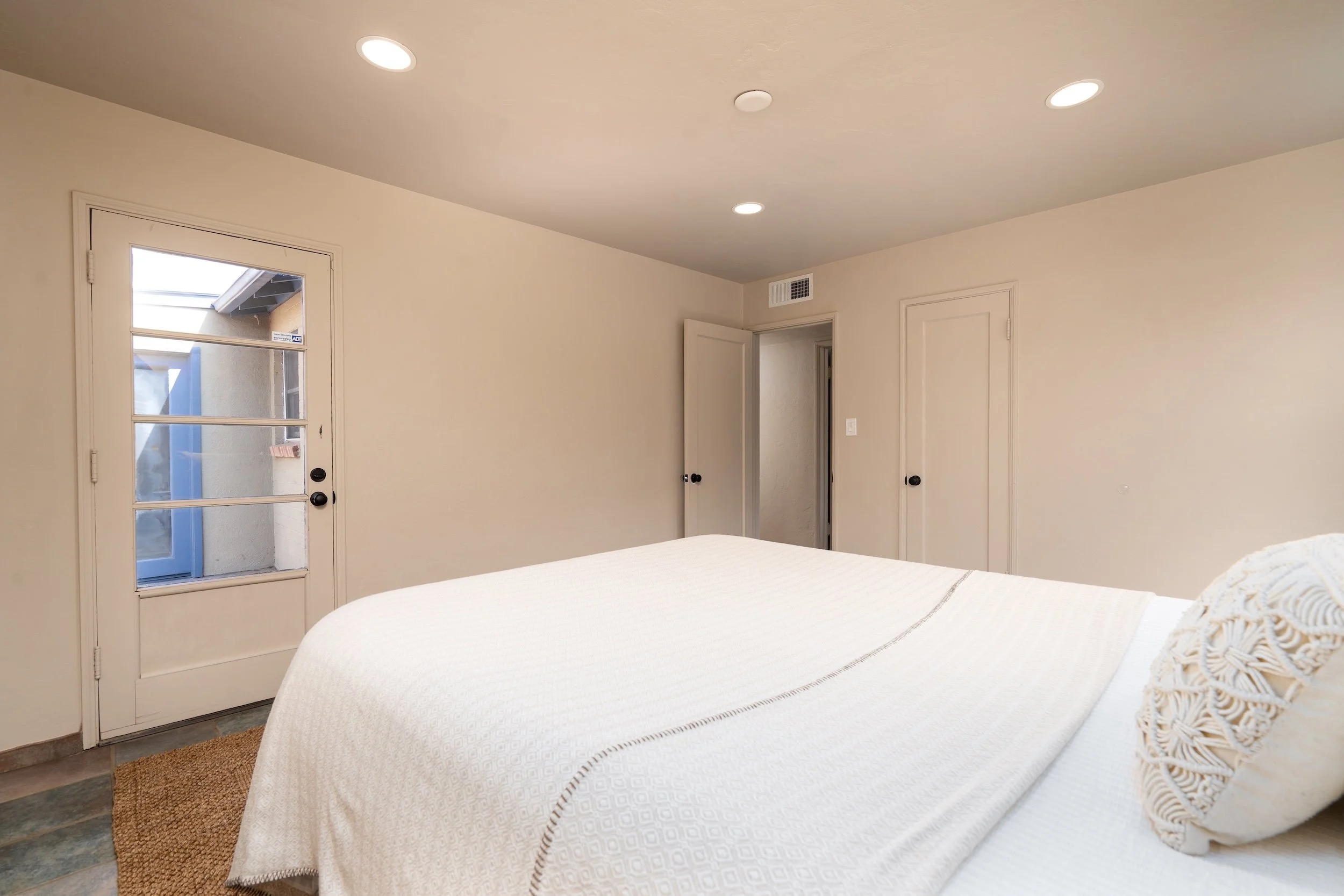 A bedroom with a white bedspread, a woven rug, a door with glass panes leading outside, and an open door to another room. Recessed ceiling lights illuminate the space.