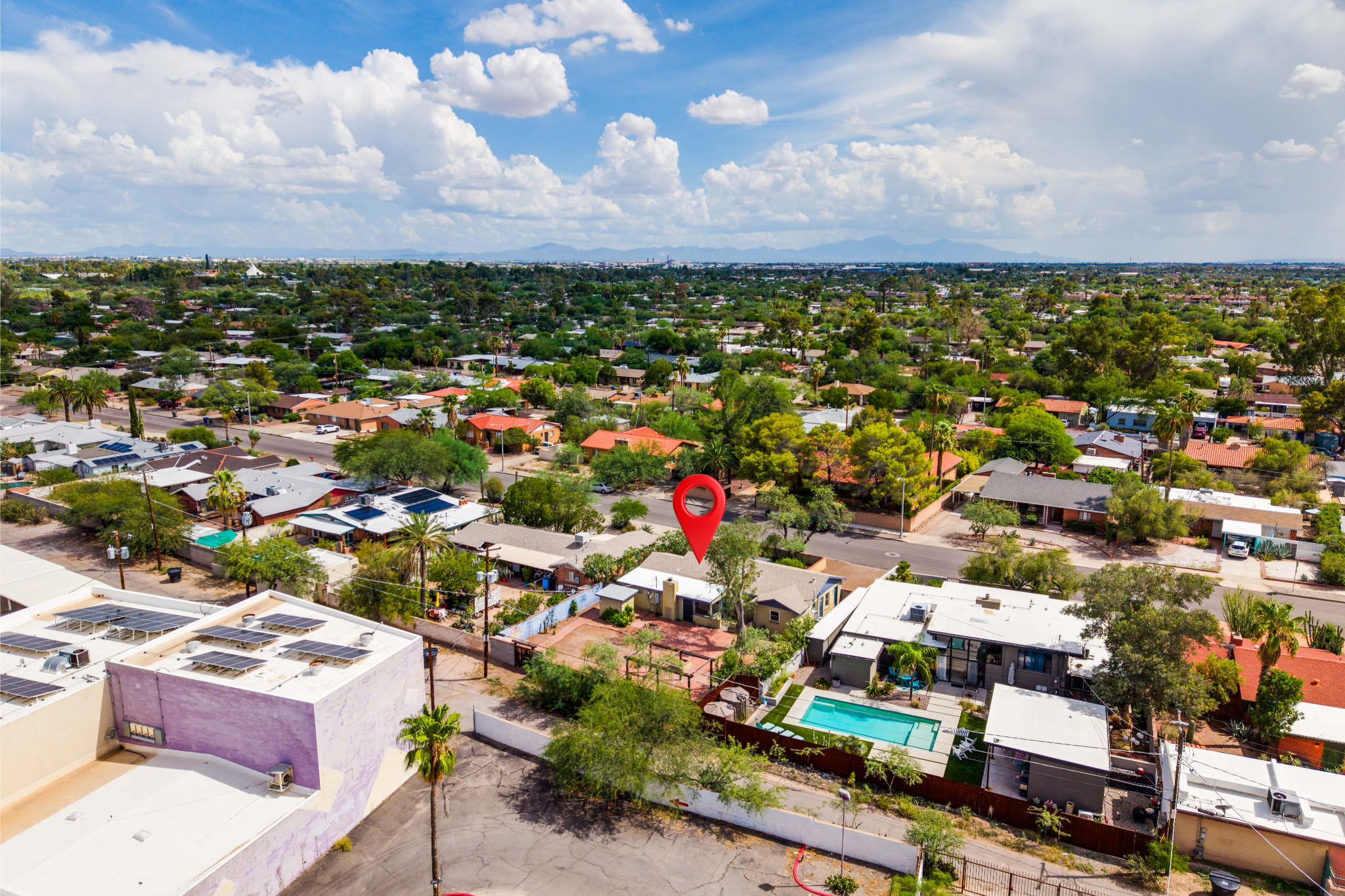 Aerial view of a suburban neighborhood with houses, trees, and a swimming pool, under a partly cloudy sky. Mountains are visible in the distance.