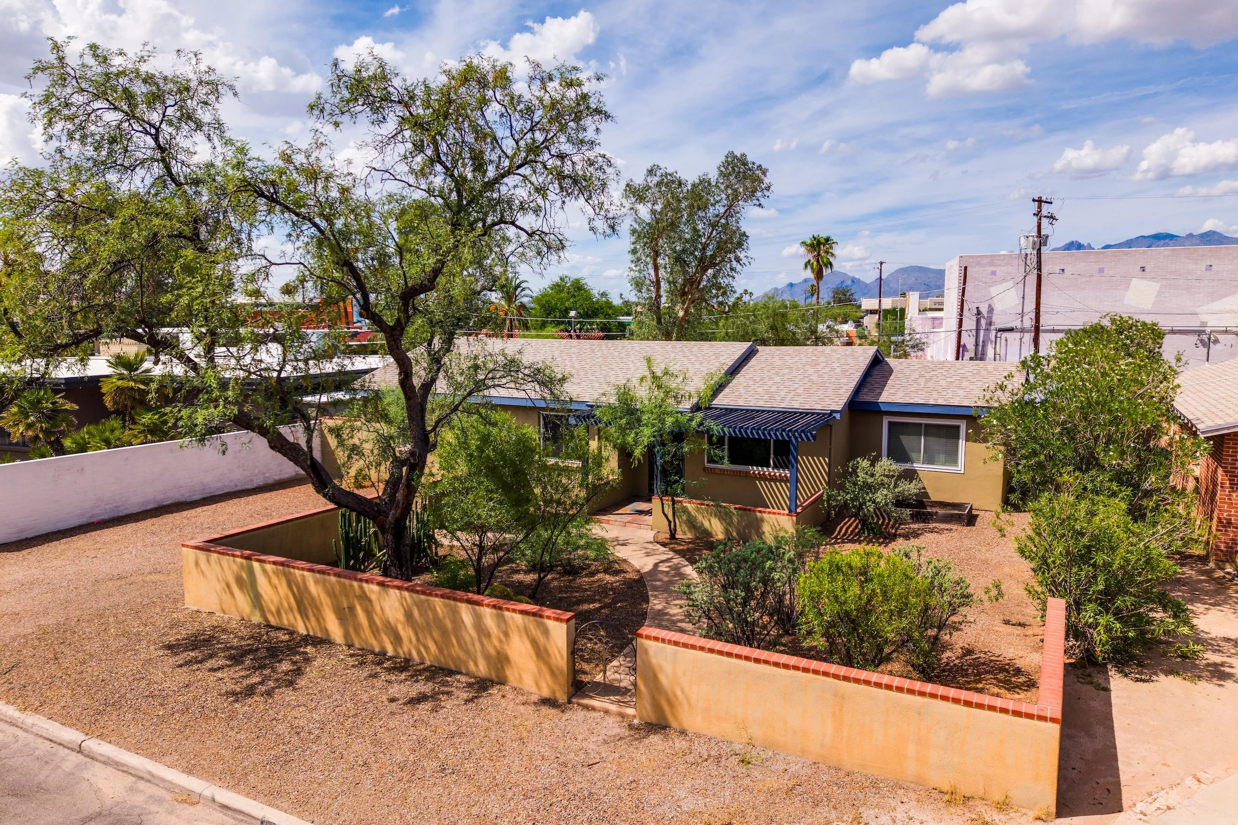 View of residential neighborhood with desert landscaping, trees, rooftops, utility poles, and distant mountains under a partly cloudy sky.