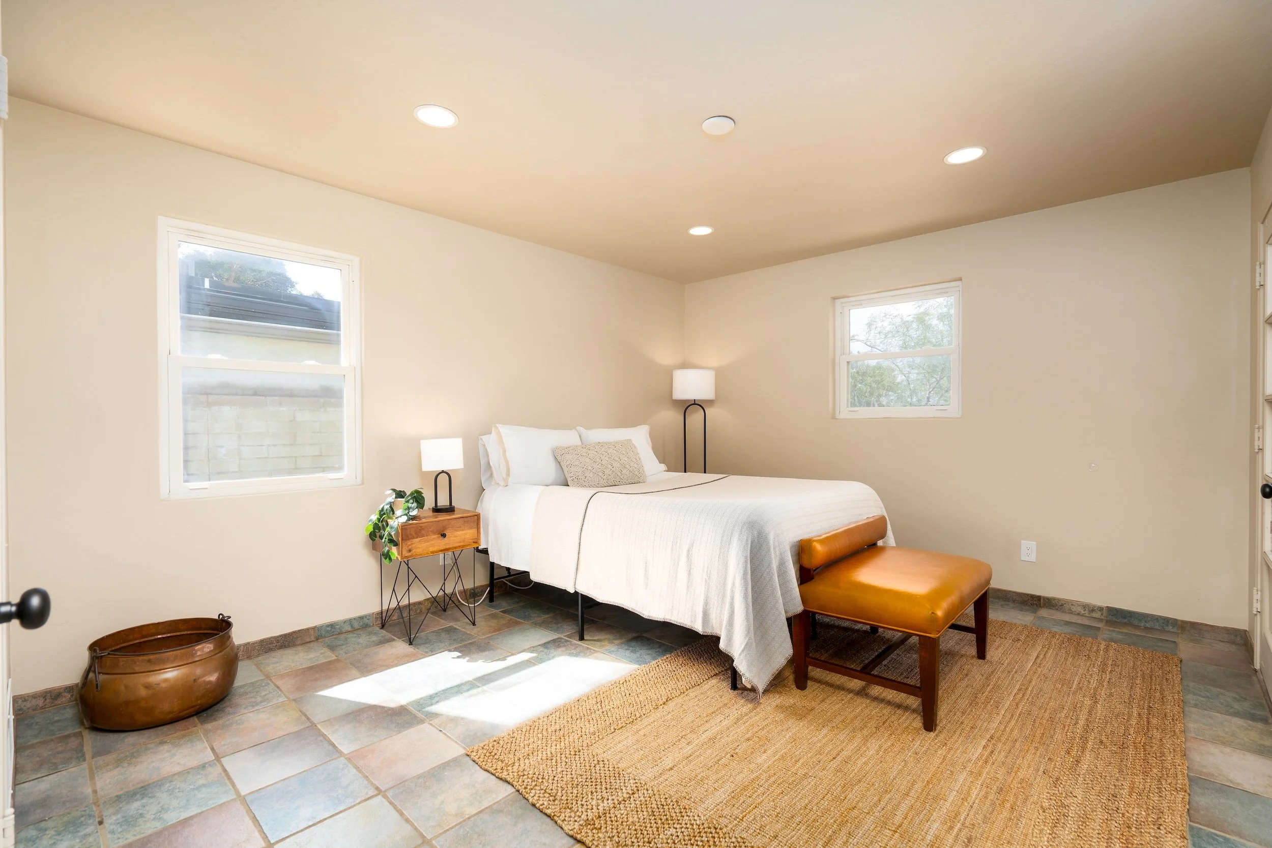 A bedroom with beige walls and two windows, a white bed with a beige pillow, a black floor lamp, a wooden bedside table with a white lamp, a leather ottoman, a large pot on the tiled floor, and a woven rug.