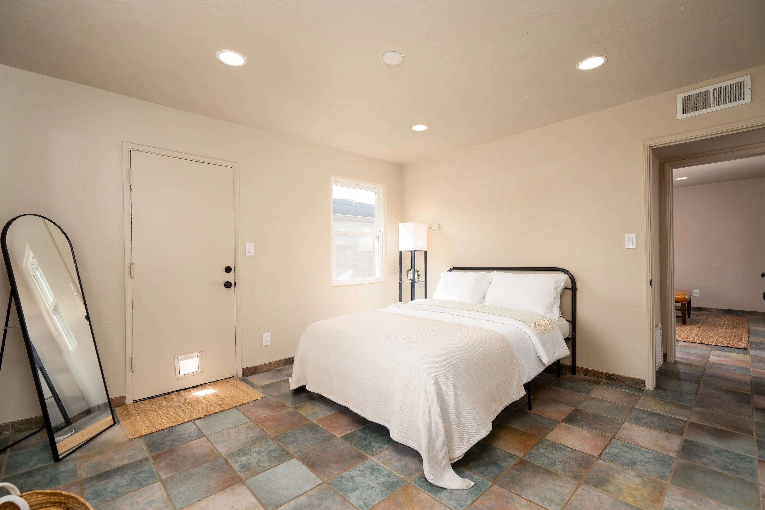 A cozy bedroom with a black metal bed frame, white bedding, and a small black shelving unit with a potted plant, illuminated by sunlight from a window. The room has beige walls, a tiled floor, and a mirror leaning against the wall near the door.