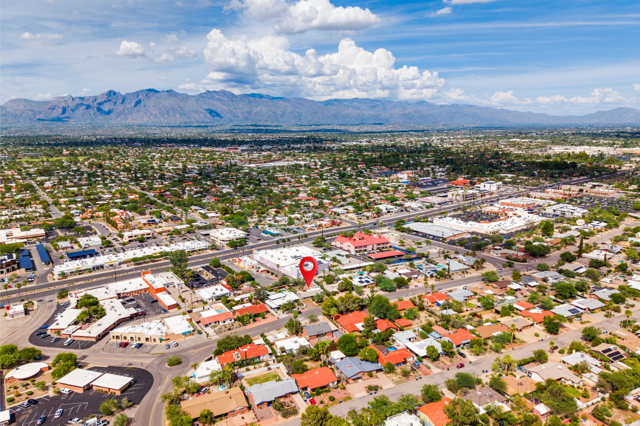 Aerial view of a city with a mountain range in the background, under a partly cloudy sky. The city includes residential houses, commercial buildings, parking lots, and streets. A red location marker is visible over a specific area.