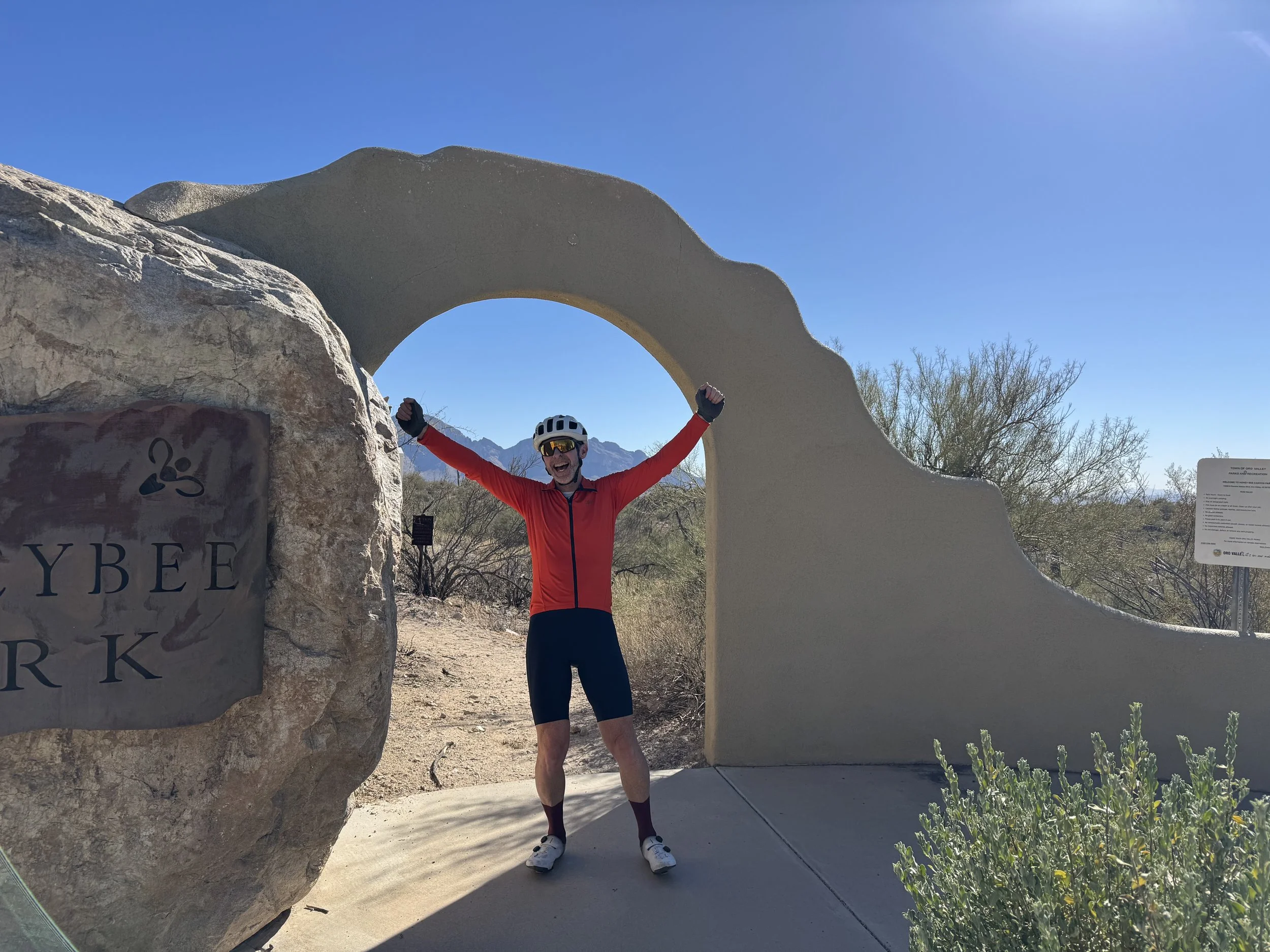 A man in red cycling gear celebrating with arms raised, standing in front of a large stone arch at HoneyBee Park during sunny weather.