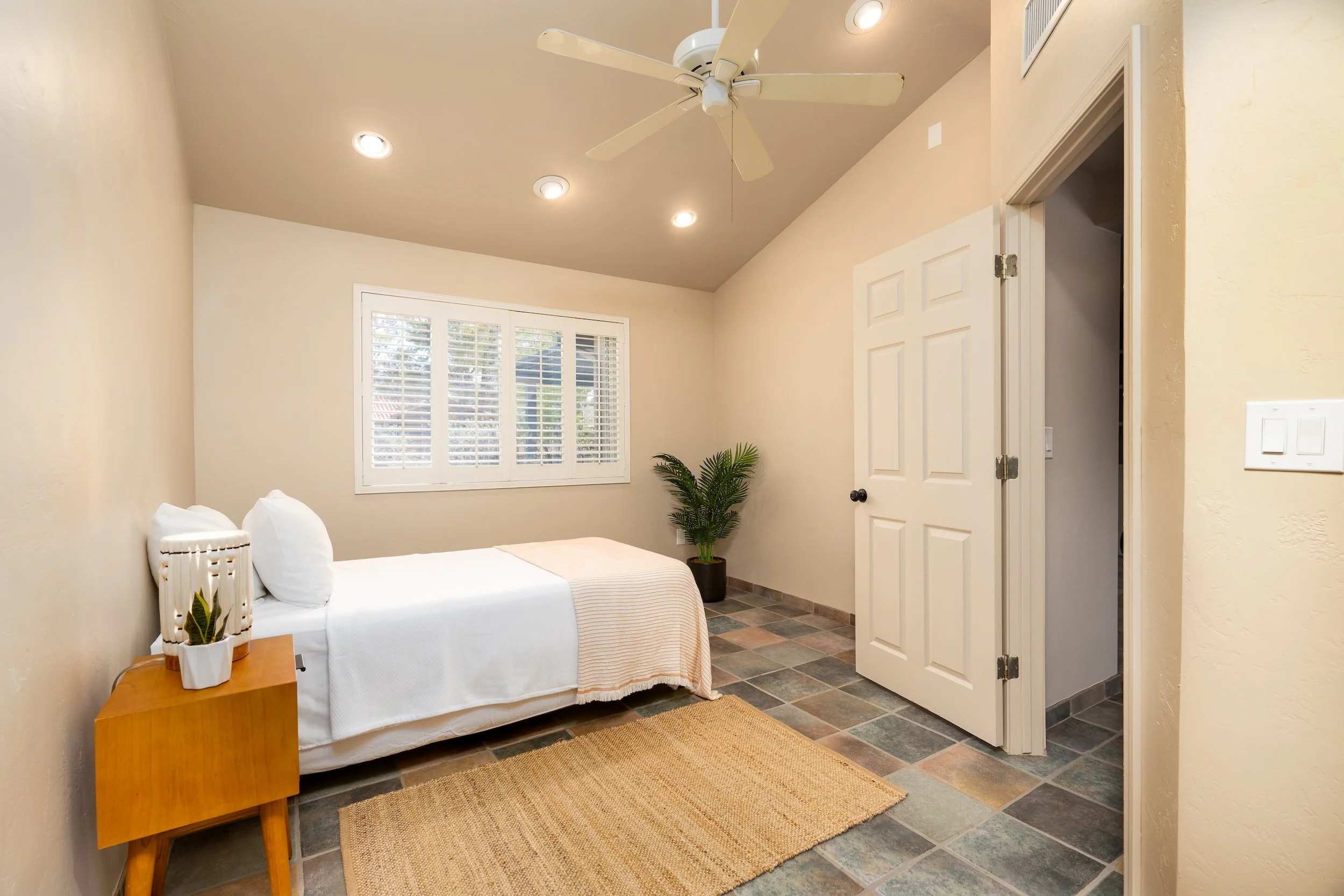 A bedroom with a white bed, a wooden side table with plants, a window with white shutters, a ceiling fan, recessed lighting, patterned tiled floor, and a closet door.