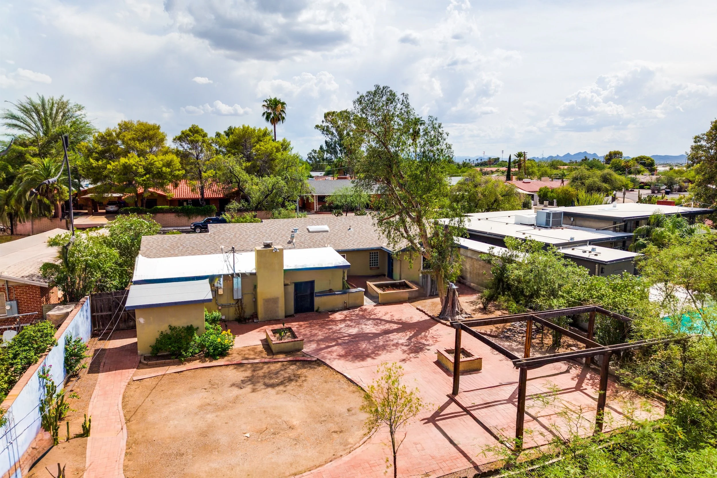 A suburban neighborhood with single-story houses, trees, and a partly cloudy sky. The foreground shows a yard with a brick pathway and a small garden, while the background features rooftops and distant mountains.