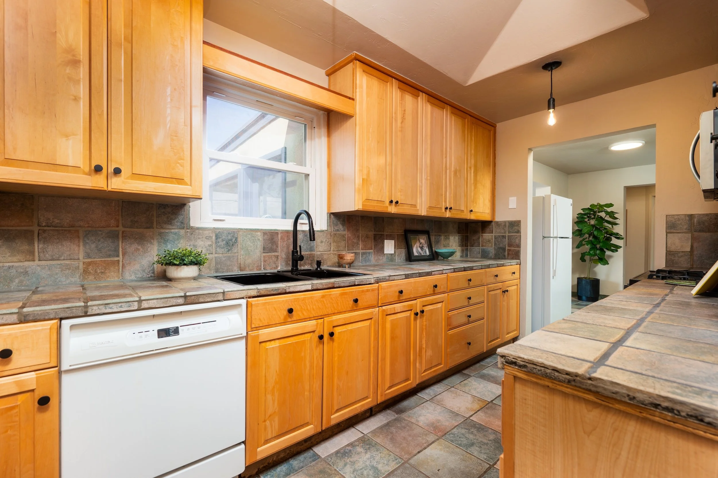 A kitchen with wooden cabinets, a tiled countertop, a window above the sink, a potted plant, a dishwasher, and a doorway leading to another room.