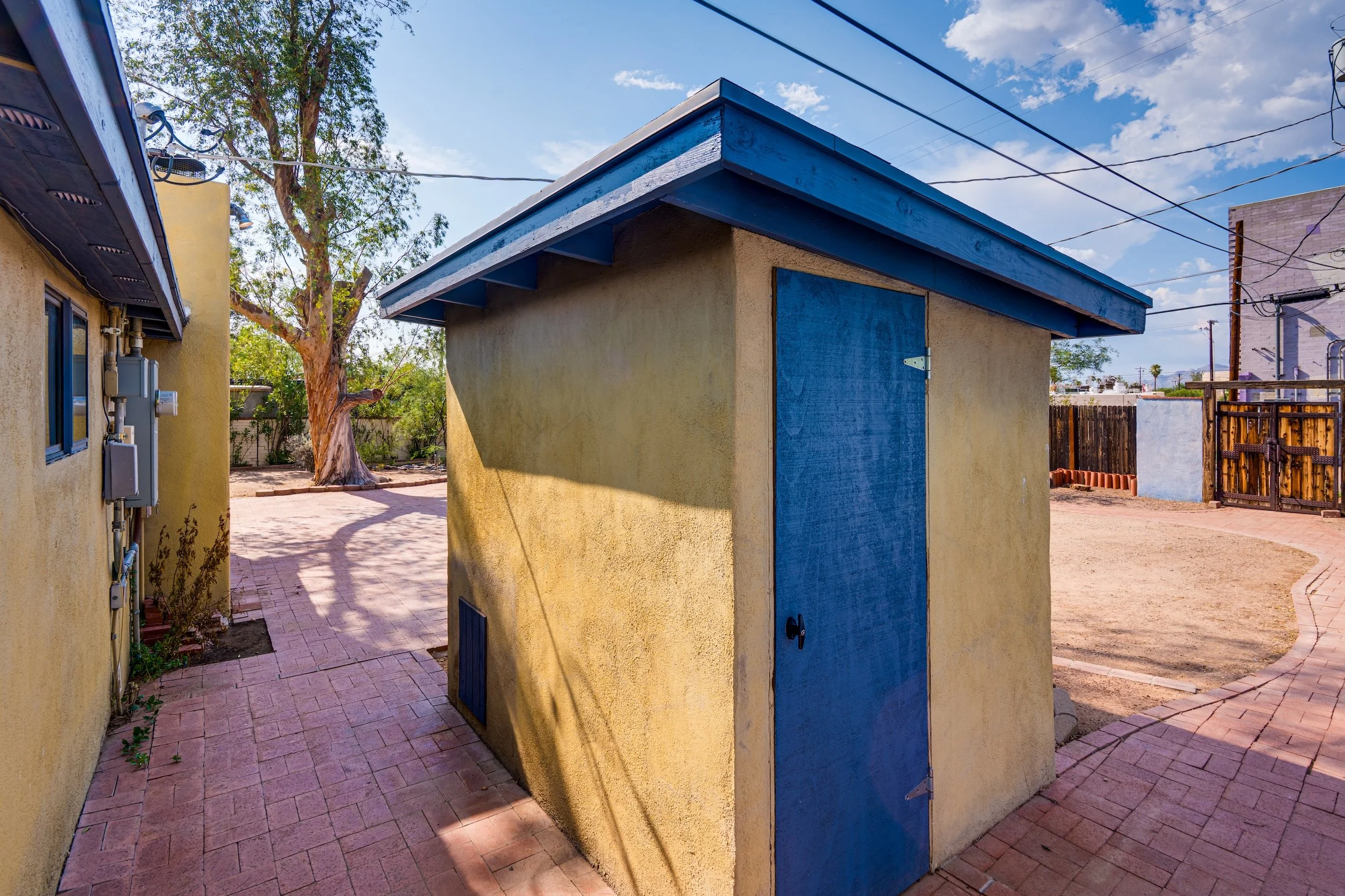 Exterior view of a small beige building with a blue roof and blue door, situated between yellow and gray neighboring structures. The setting includes a tree, electrical wires, and a dirt area under a partly cloudy sky.