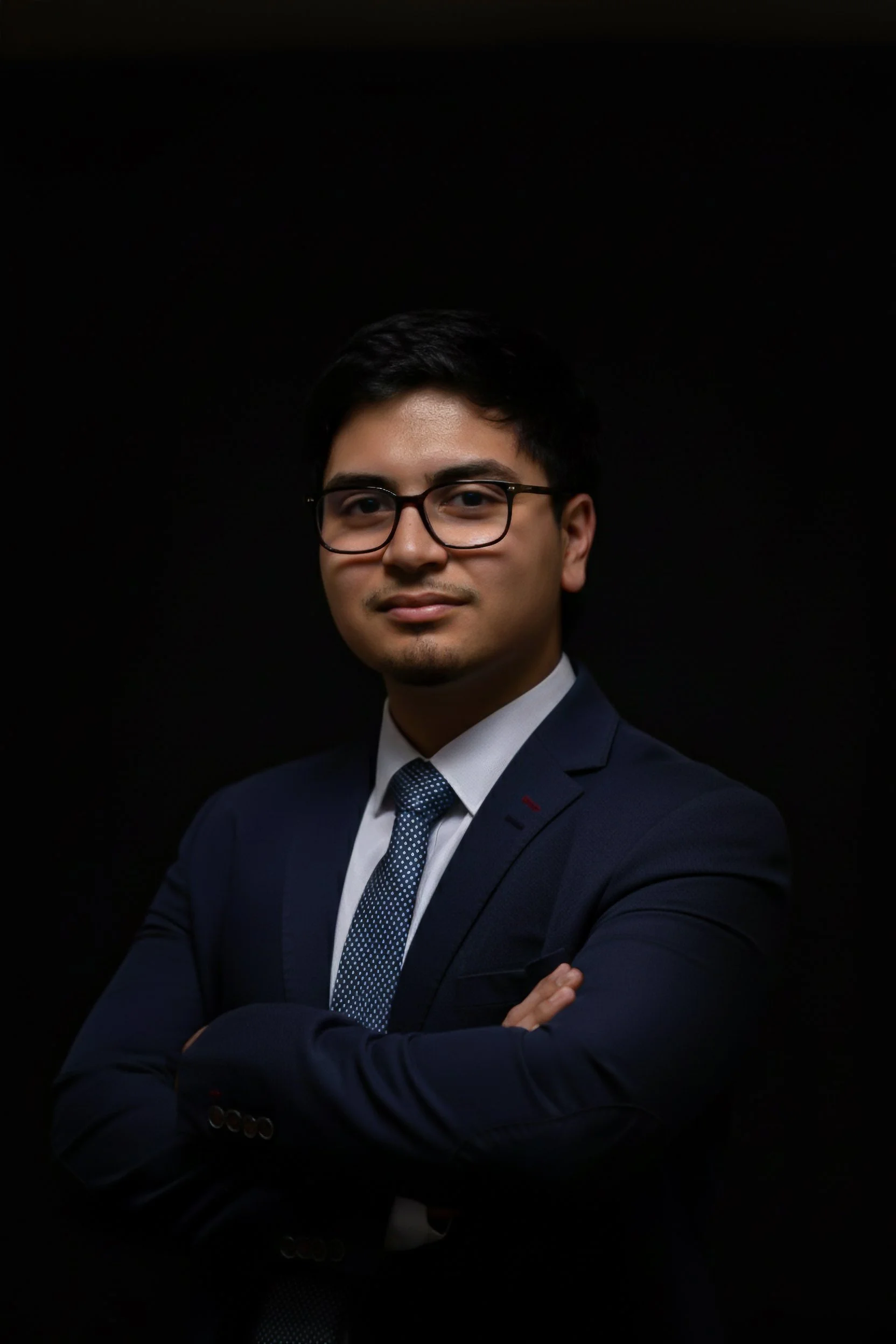 A young man with black hair, wearing glasses and a navy blue suit with a white shirt and a blue patterned tie, is posing with his arms crossed against a black background.