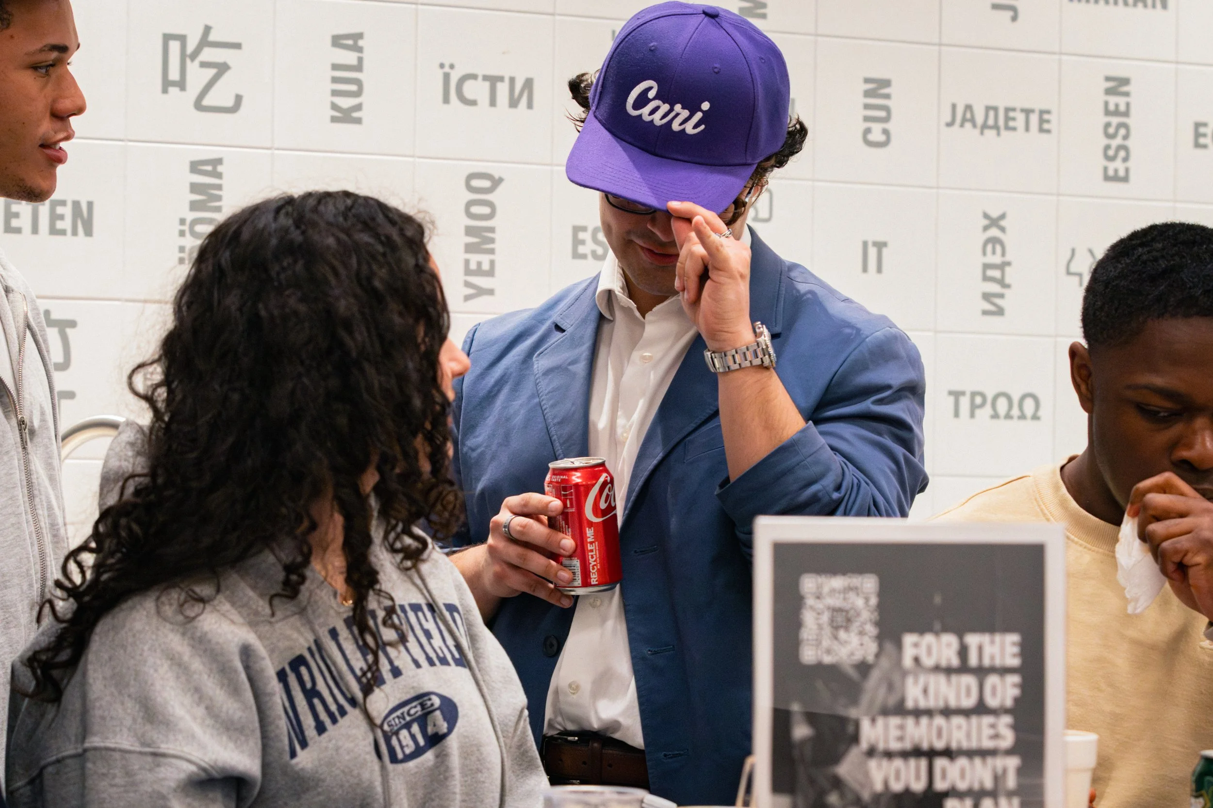 A man wearing glasses, a blue jacket, and a purple cap with the name 'Cari' is holding a can of Coca-Cola. He is surrounded by a group of people, and they are indoors against a white tiled wall with words in various languages. The man appears to be l