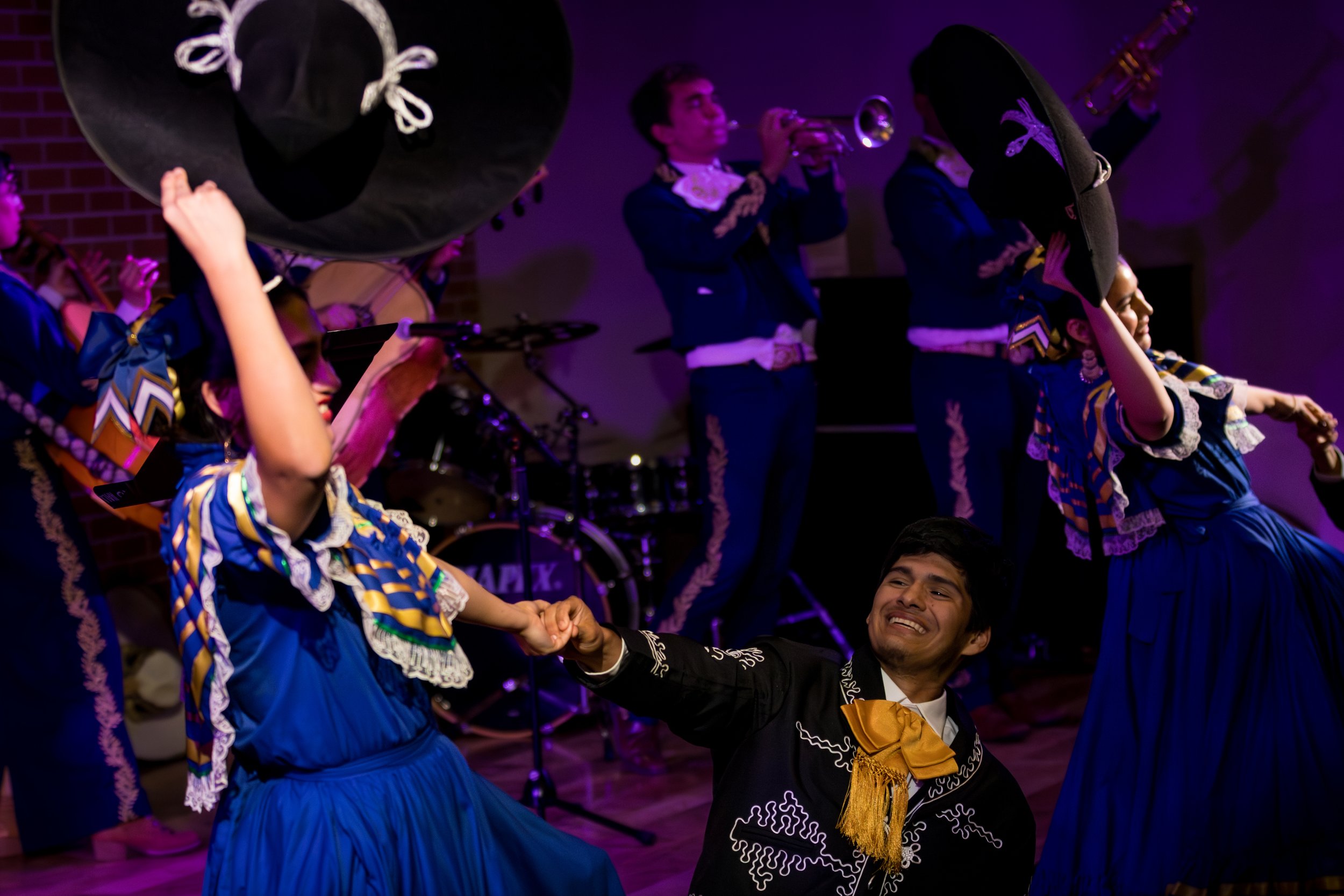 People performing traditional Mexican folk dance, wearing colorful costumes, on stage with one person playing trumpet, in a lively setting.