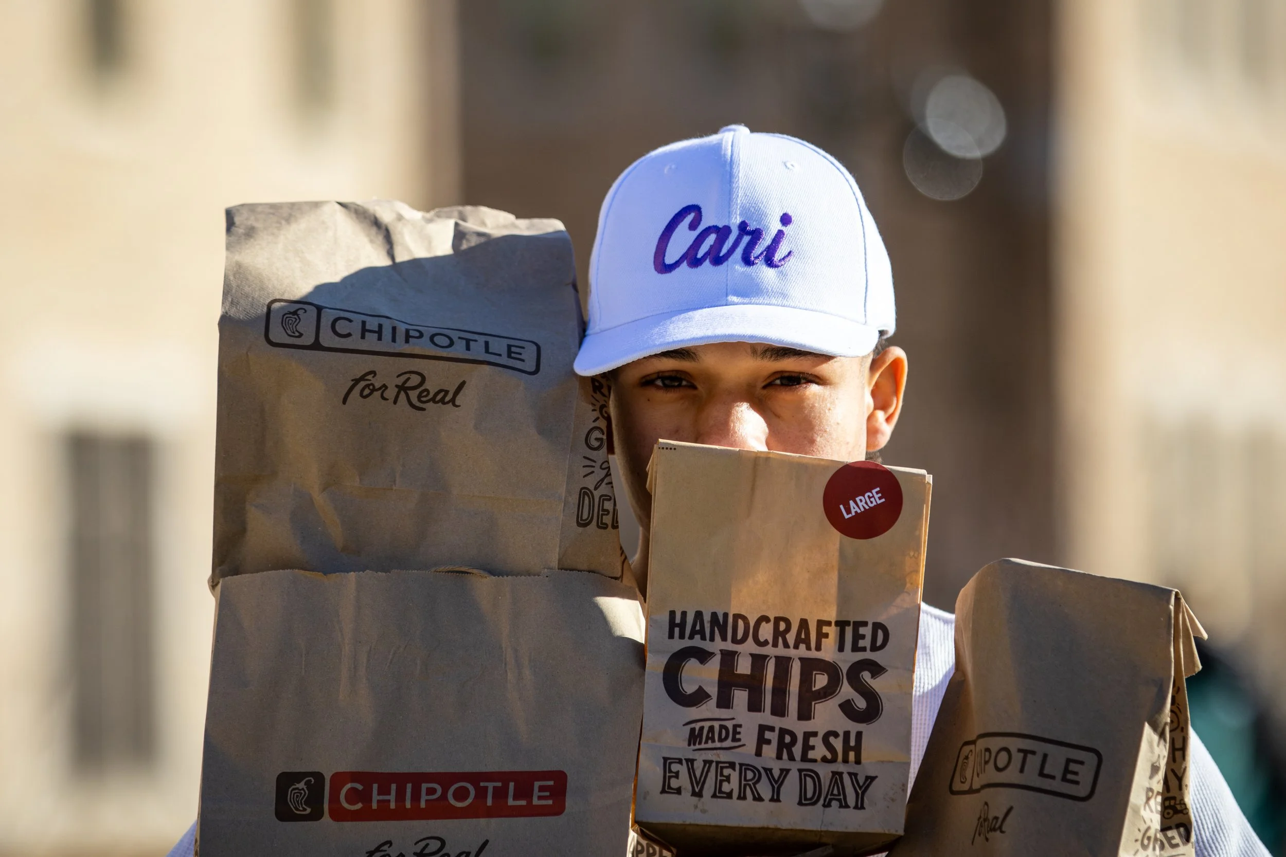 A young boy wearing a white cap embroidered with 'Cari' holding a large paper bag from Chipotle Mexican Grill, with other paper bags from Chipotle visible around him, outdoors on a sunny day.
