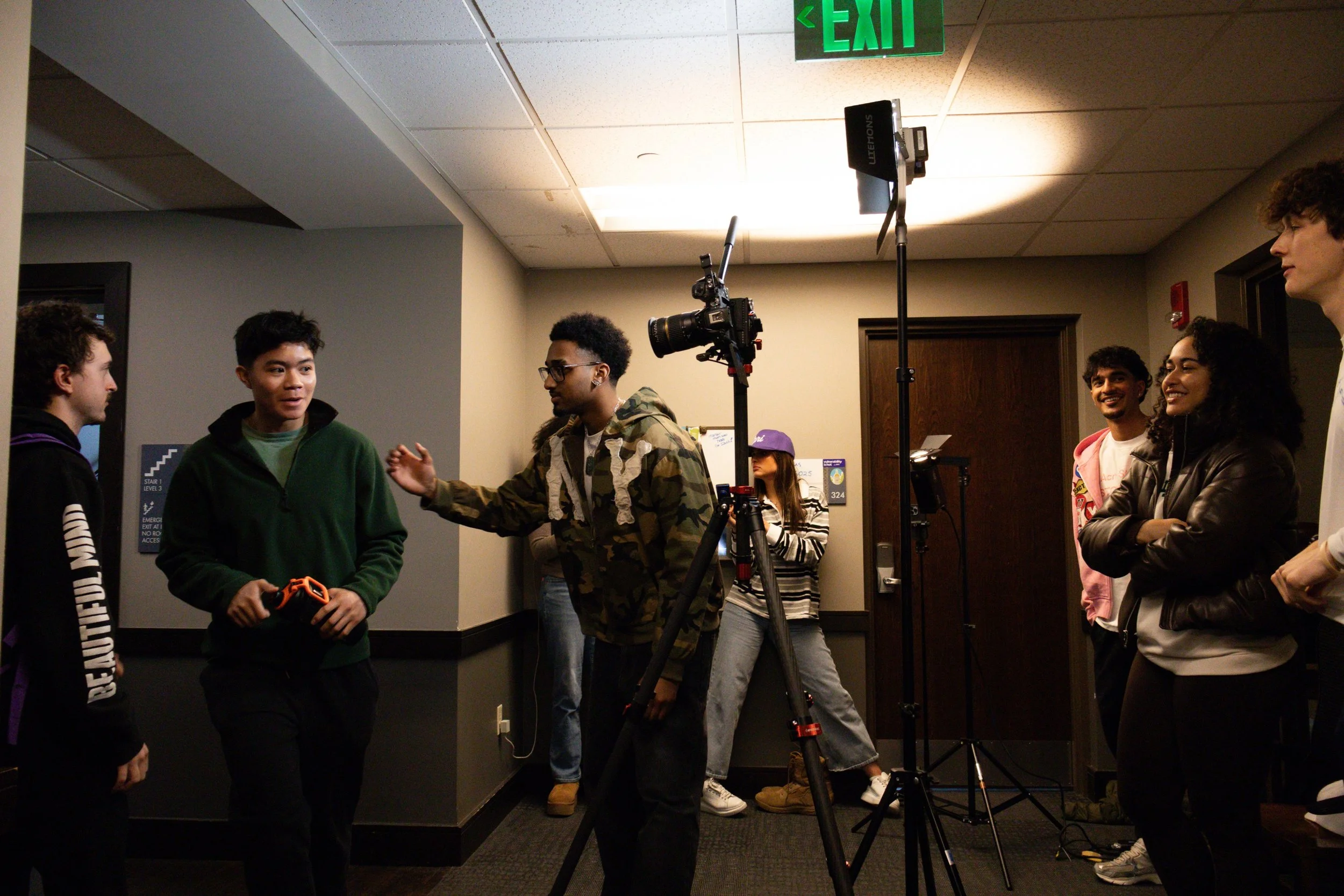 Group of young people setting up filming equipment in an indoor hallway, with a green exit sign on the ceiling.