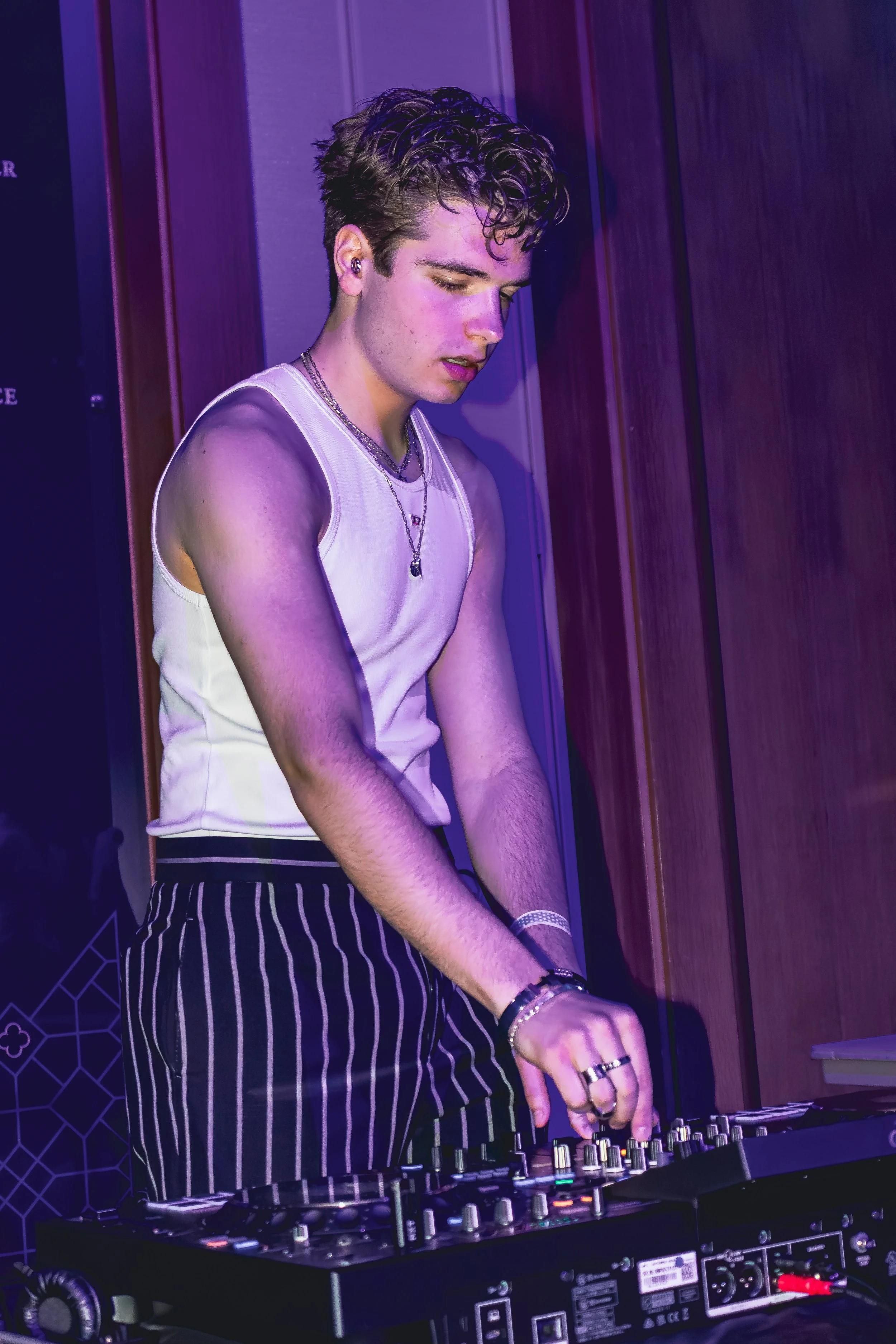 A young man with curly hair wearing a white sleeveless shirt and striped pants, DJing with headphones and a mixer in a dimly lit room with purple lighting.