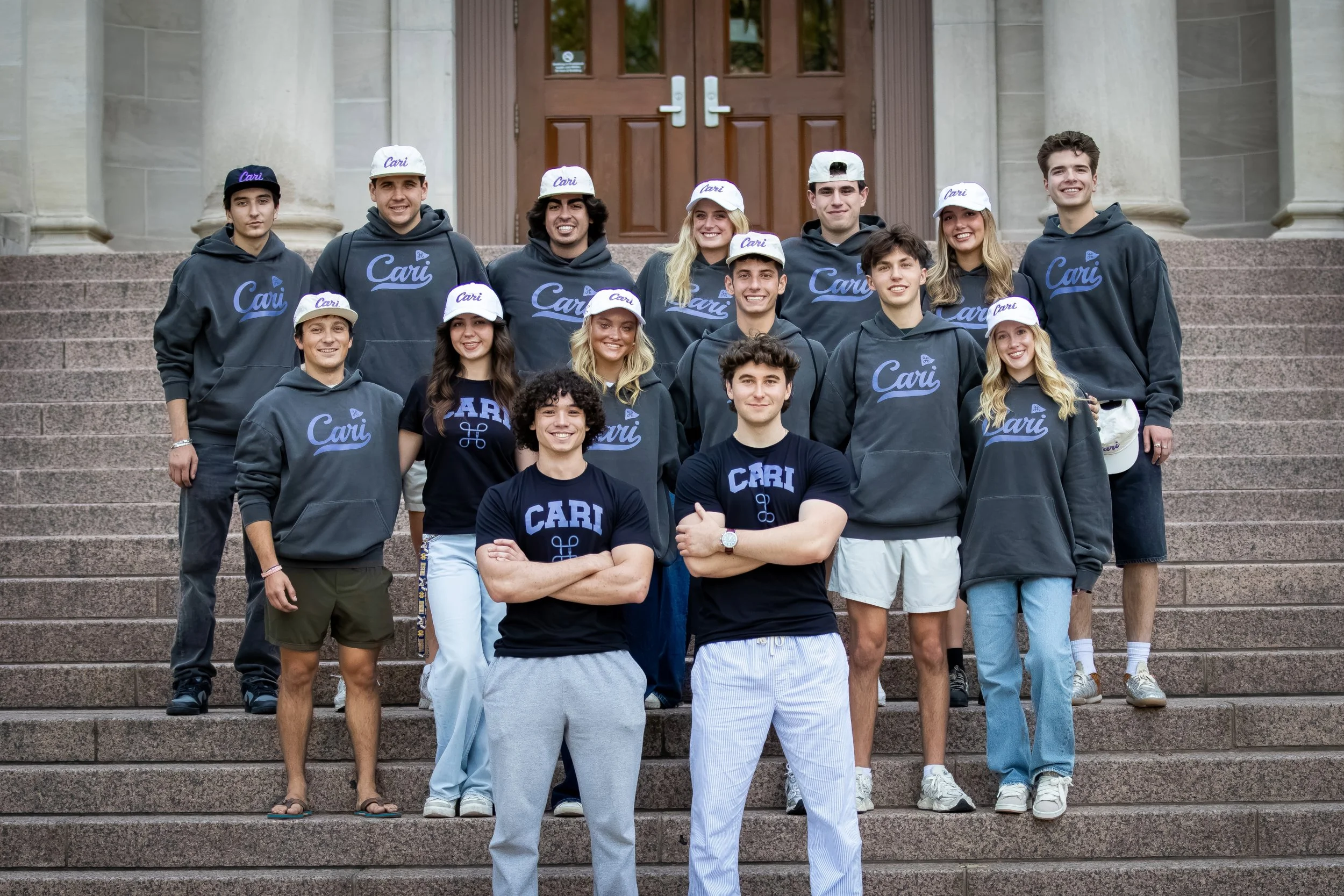 Group of young people standing on stone steps outside a building with large columns, wearing matching dark gray hoodies and white or dark caps with the word 'Cari' on them, smiling for a photo.