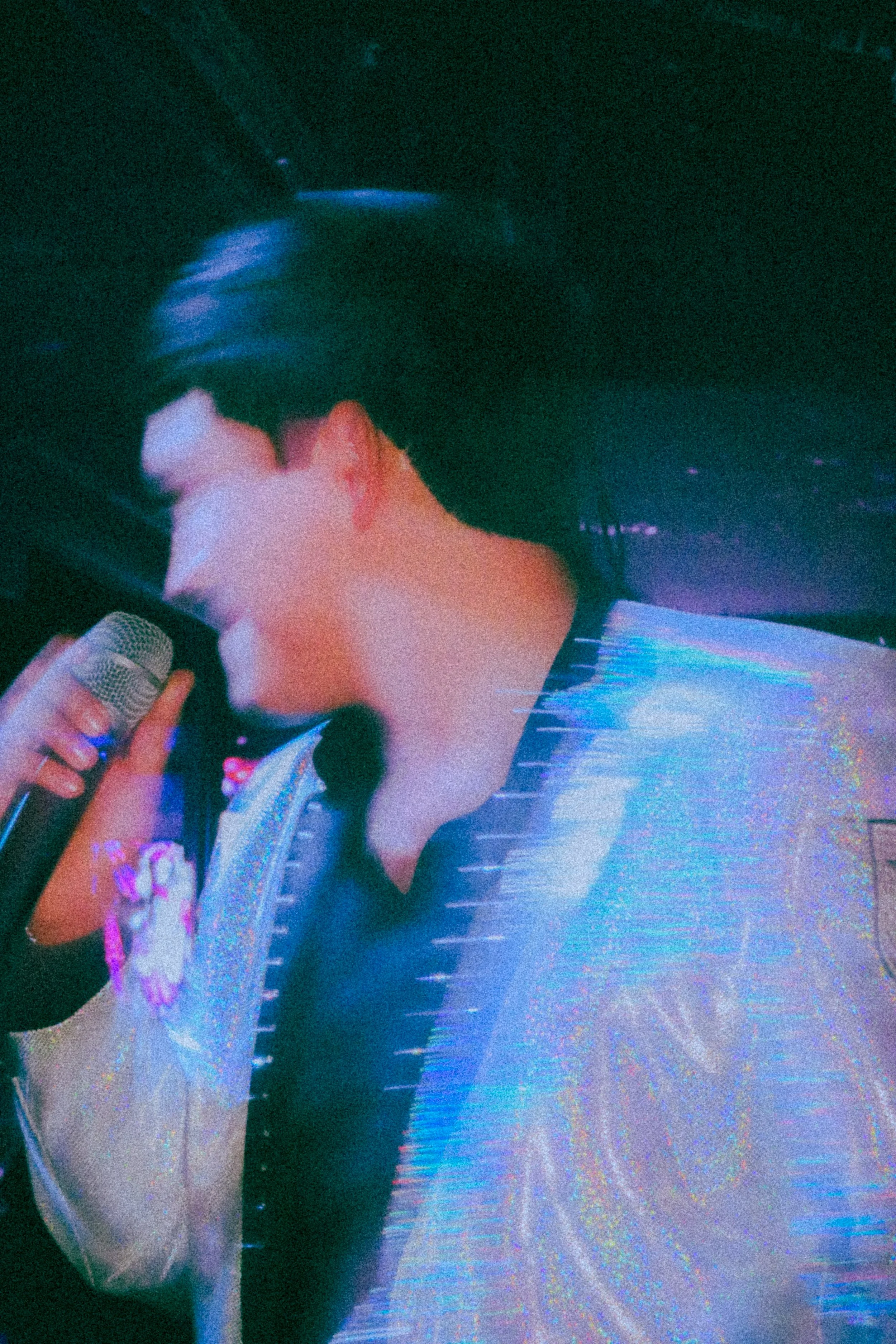 A person singing into a microphone at a dimly lit event with colorful lighting effects.