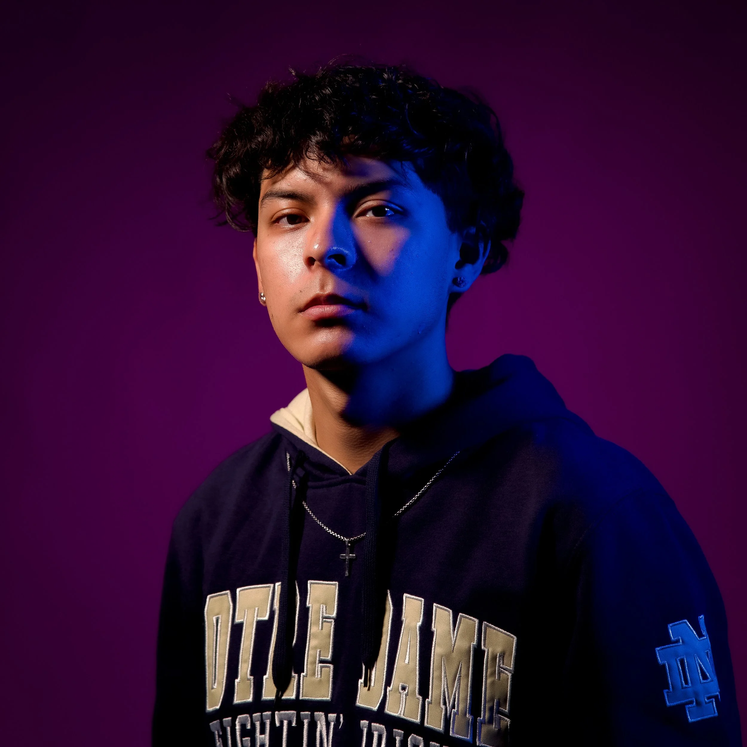 Close-up portrait of a person with curly dark hair, wearing a dark hoodie with a graphic design, standing against a purple background.