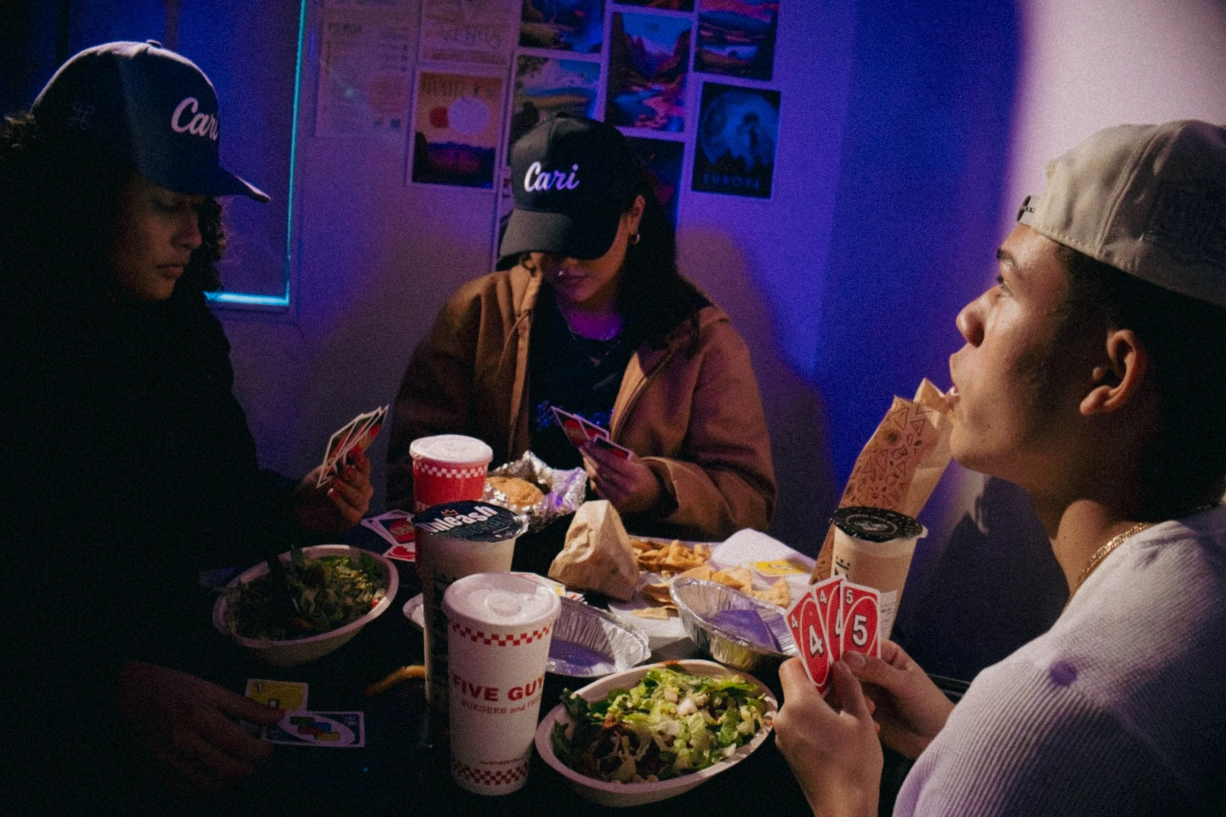 Group of people sitting around a table playing cards, with fast food items like salads, taco chips, drinks, and paper cups, in a dimly lit room with colorful posters on the wall.