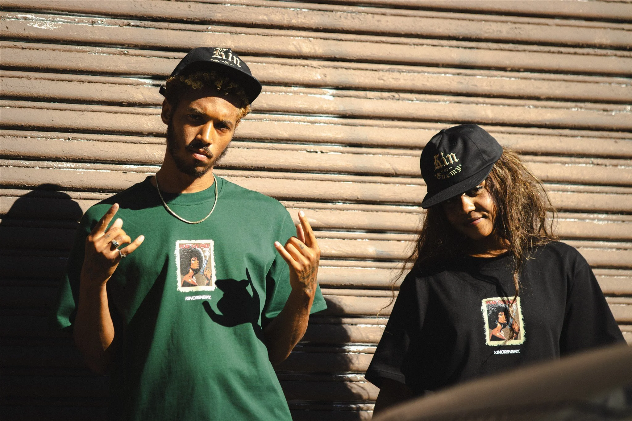Two young adults standing in front of a wooden roller shutter, both wearing black hats with text and graphic T-shirts with printed images of a woman. The person on the left is a young man with curly hair, a mustache, and goatee, making a hand gesture