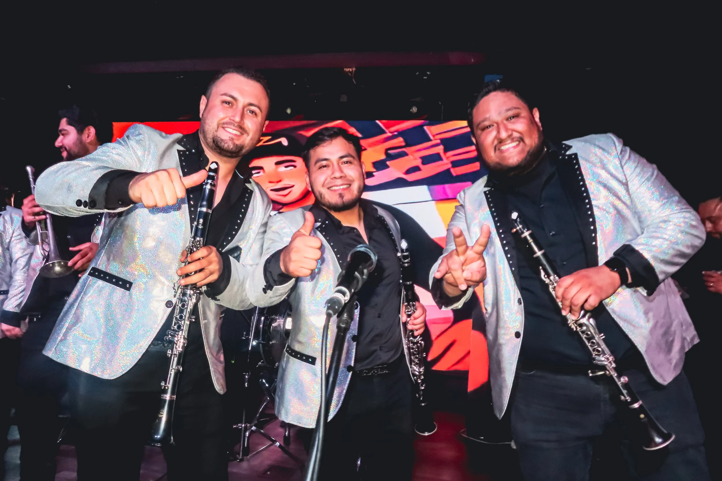 Three musicians in shiny silver jackets holding clarinets, smiling, posing on stage with a red and black background.
