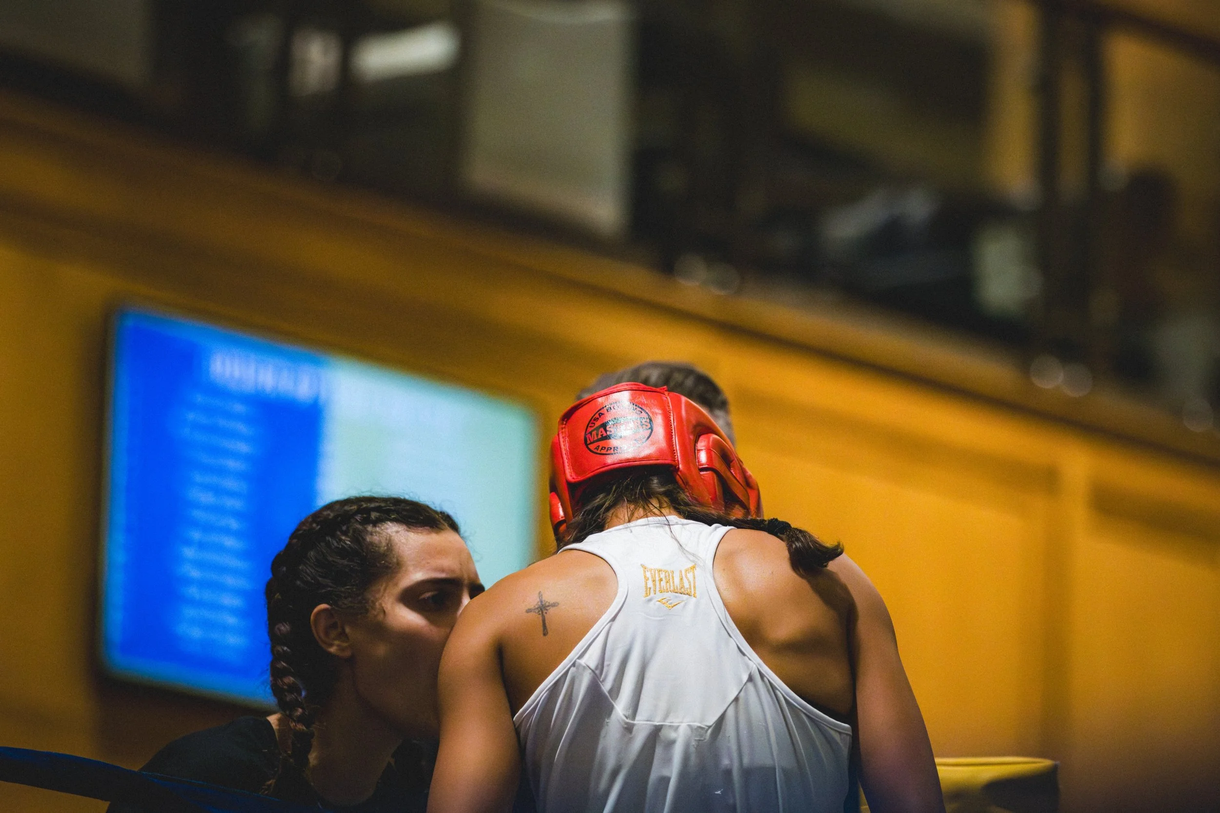 Two women, one of whom is wearing a red headgear, embrace in a boxing ring. The woman in the white outfit has a small cross tattoo on her shoulder. The scene appears emotional and intense, with a yellow venue background and a blurred digital screen.