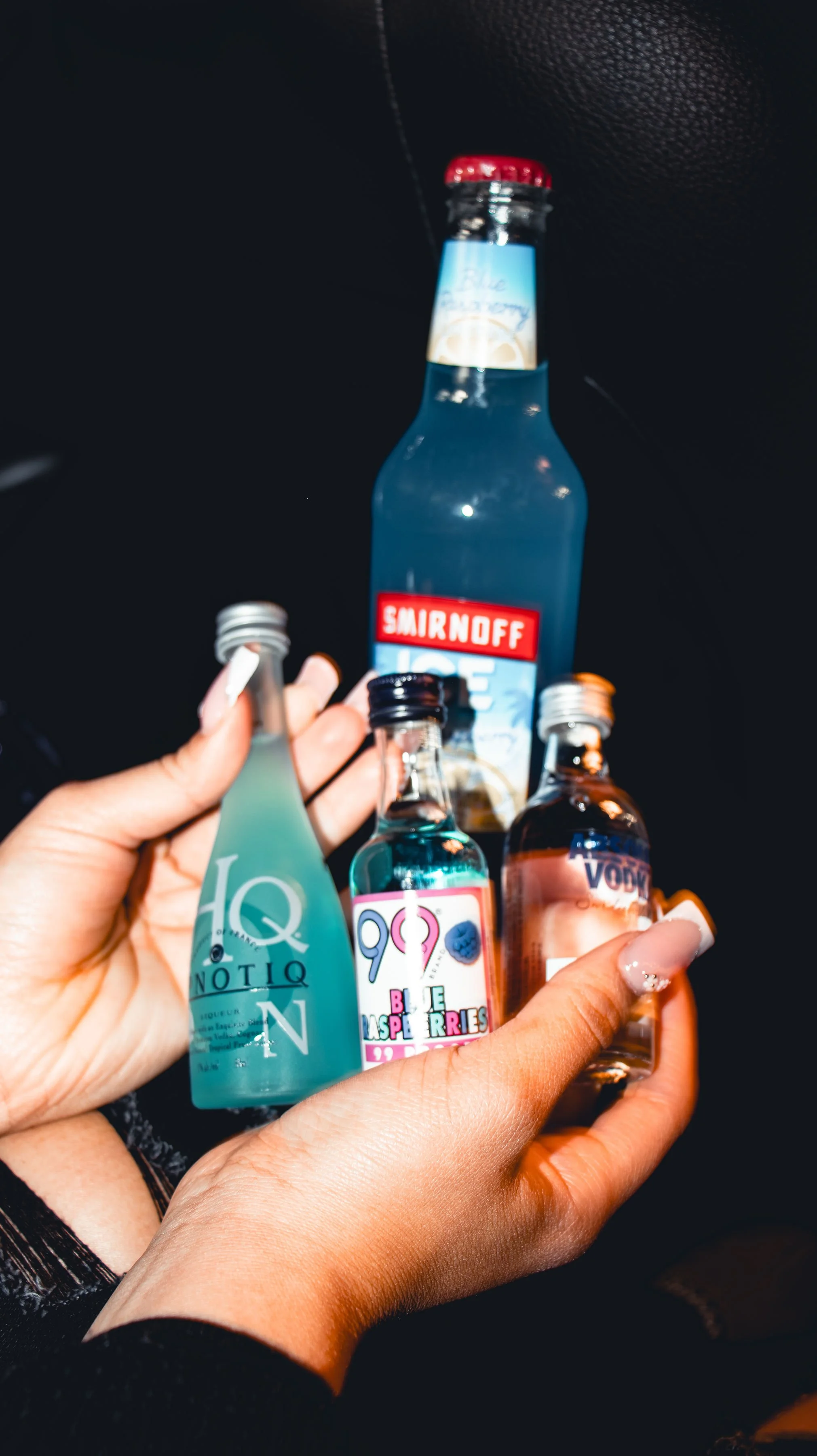 A person's hand holding four small bottles of alcohol in front of a large Smirnoff vodka bottle on a black surface.
