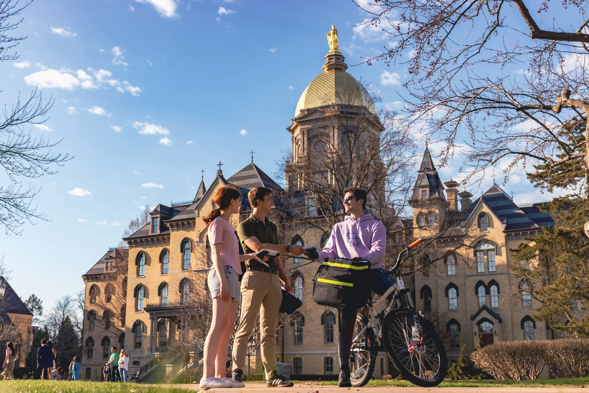 Three young people standing outside on a sunny day, two women and one man, with a historic building with a gold dome and spires in the background. One woman is holding a clipboard, the man has a bicycle, and they are engaged in conversation.