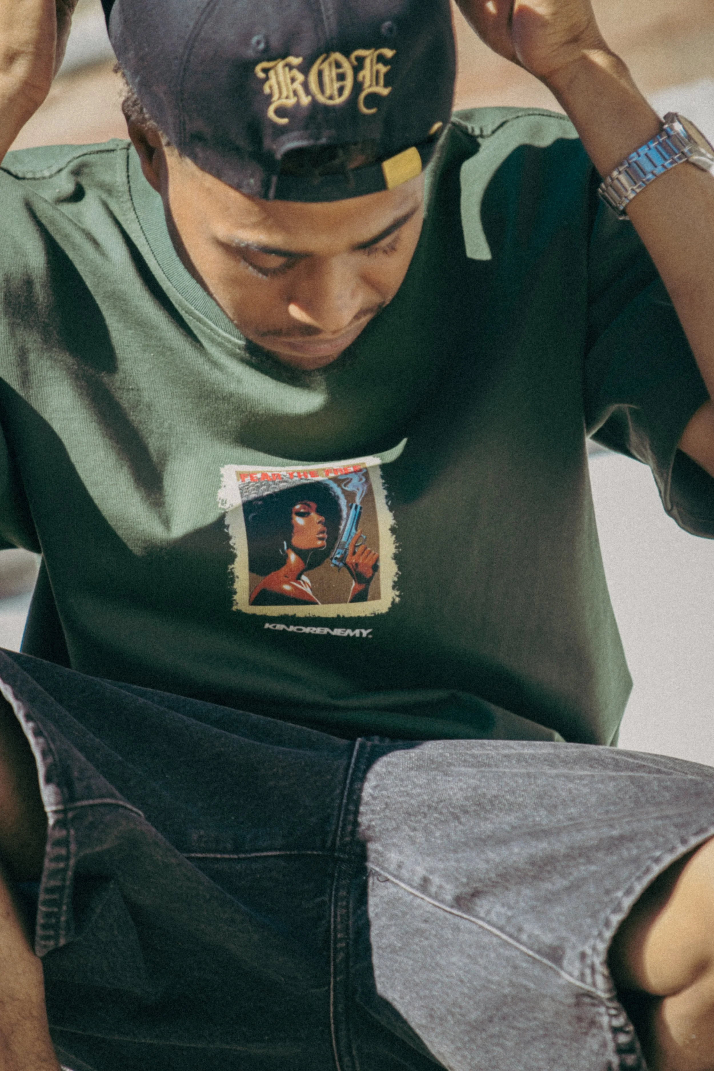A person wearing a black baseball cap with gold lettering, a green graphic T-shirt, and dark jeans, sitting outdoors.